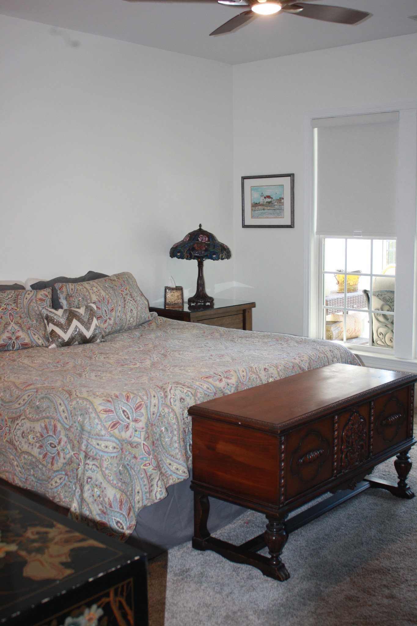 Bedroom with a pastel paisley bedspread, ornate wooden chest, and window overlooking a patio.