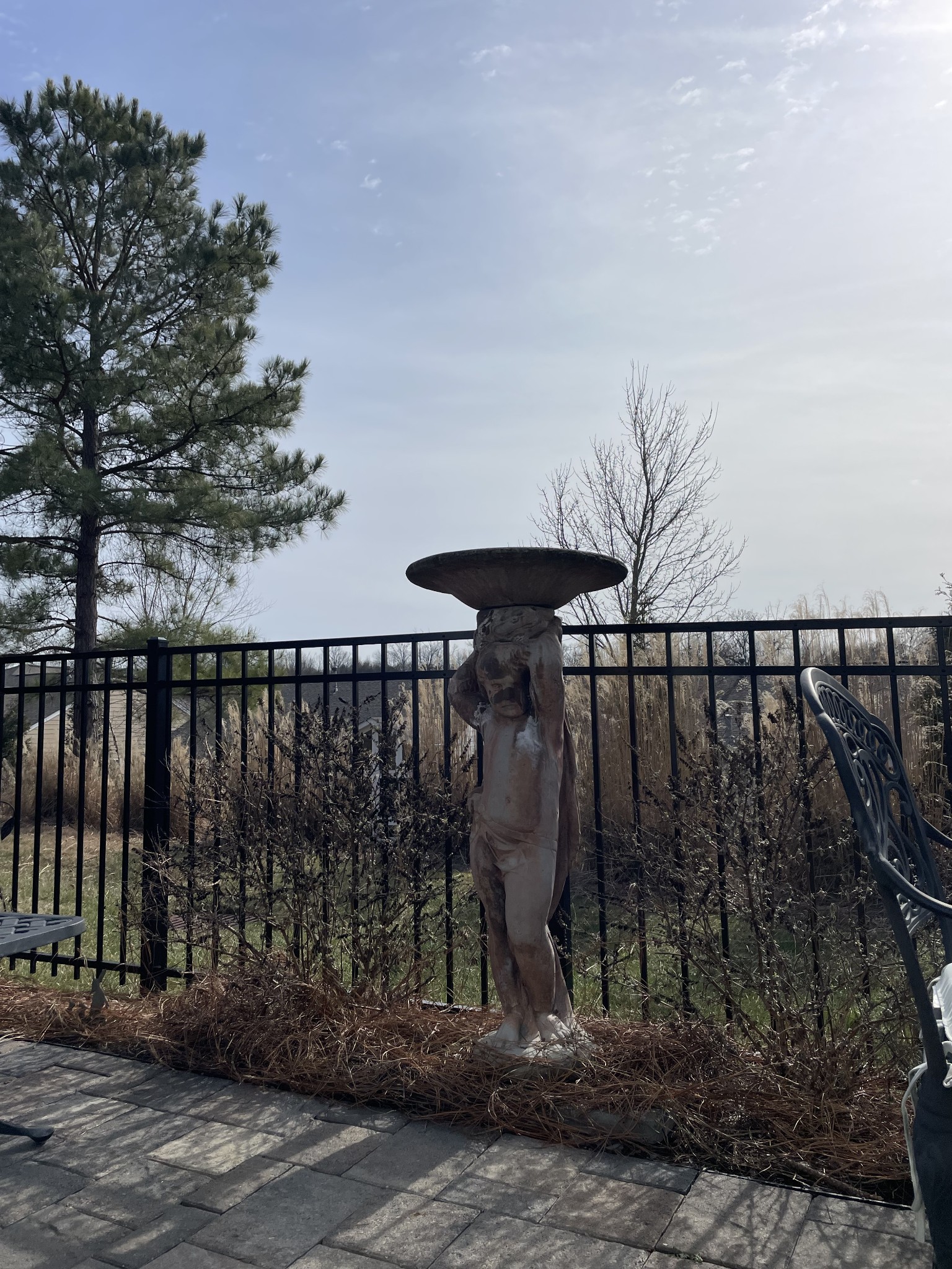 Patio with a sculpted figure supporting a wide basin beside a black metal fence and dormant shrubs.