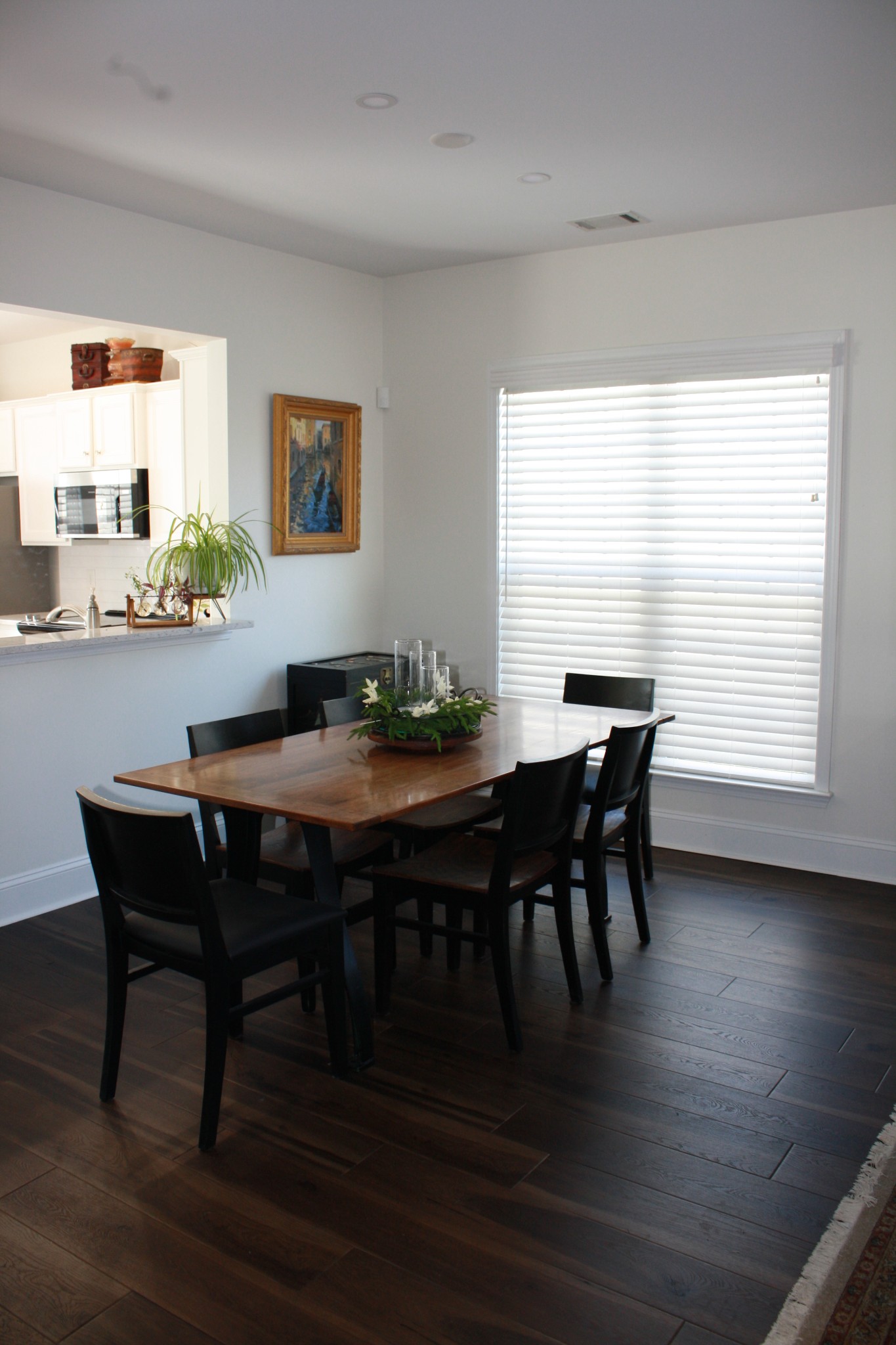 Dining room with a wooden table, dark chairs, hardwood floors, and a large window with white blinds.