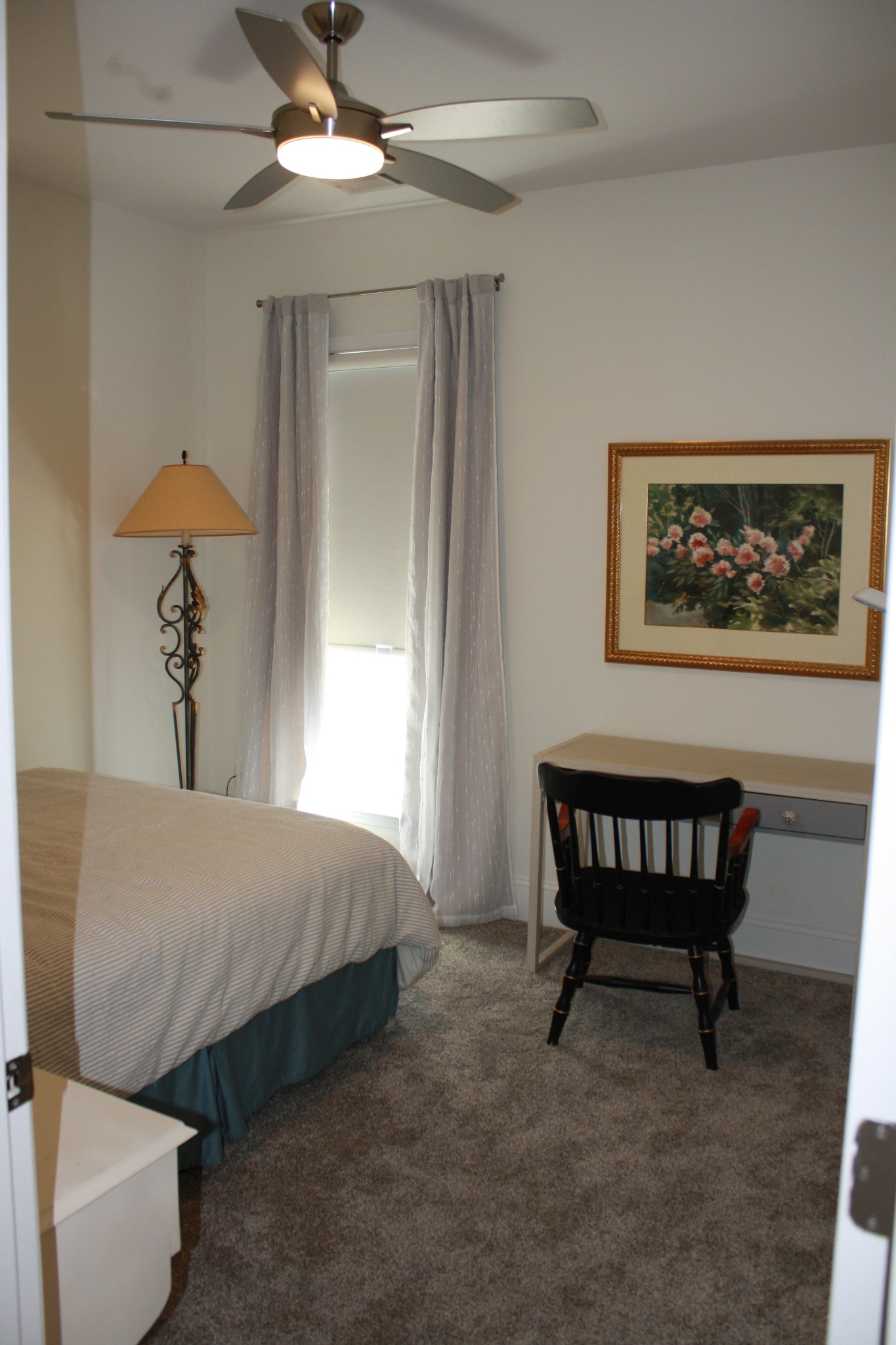 Furnished bedroom with a ceiling fan, desk, and tall window framed by sheer curtains.