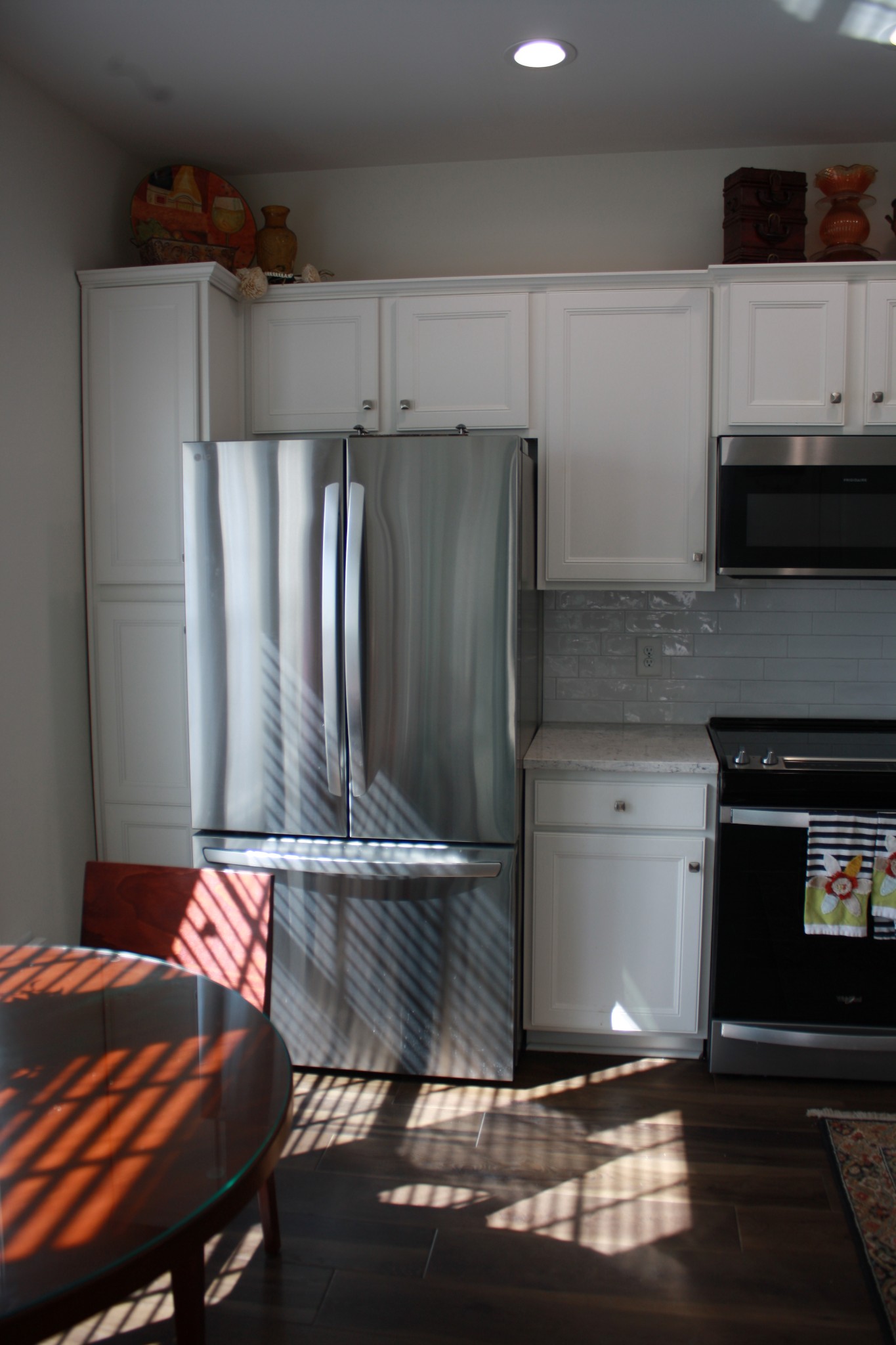 Kitchen with white cabinets and stainless steel appliances, striped sunlight falling across a glass-top table and floor.