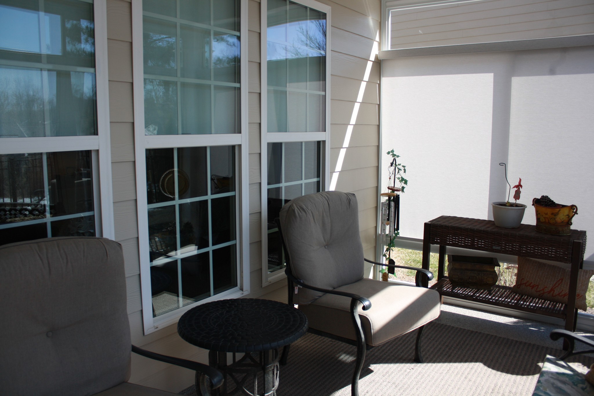 Enclosed sunroom with tall grid-pane windows, gray cushioned chairs, and a wicker console table with potted plants.