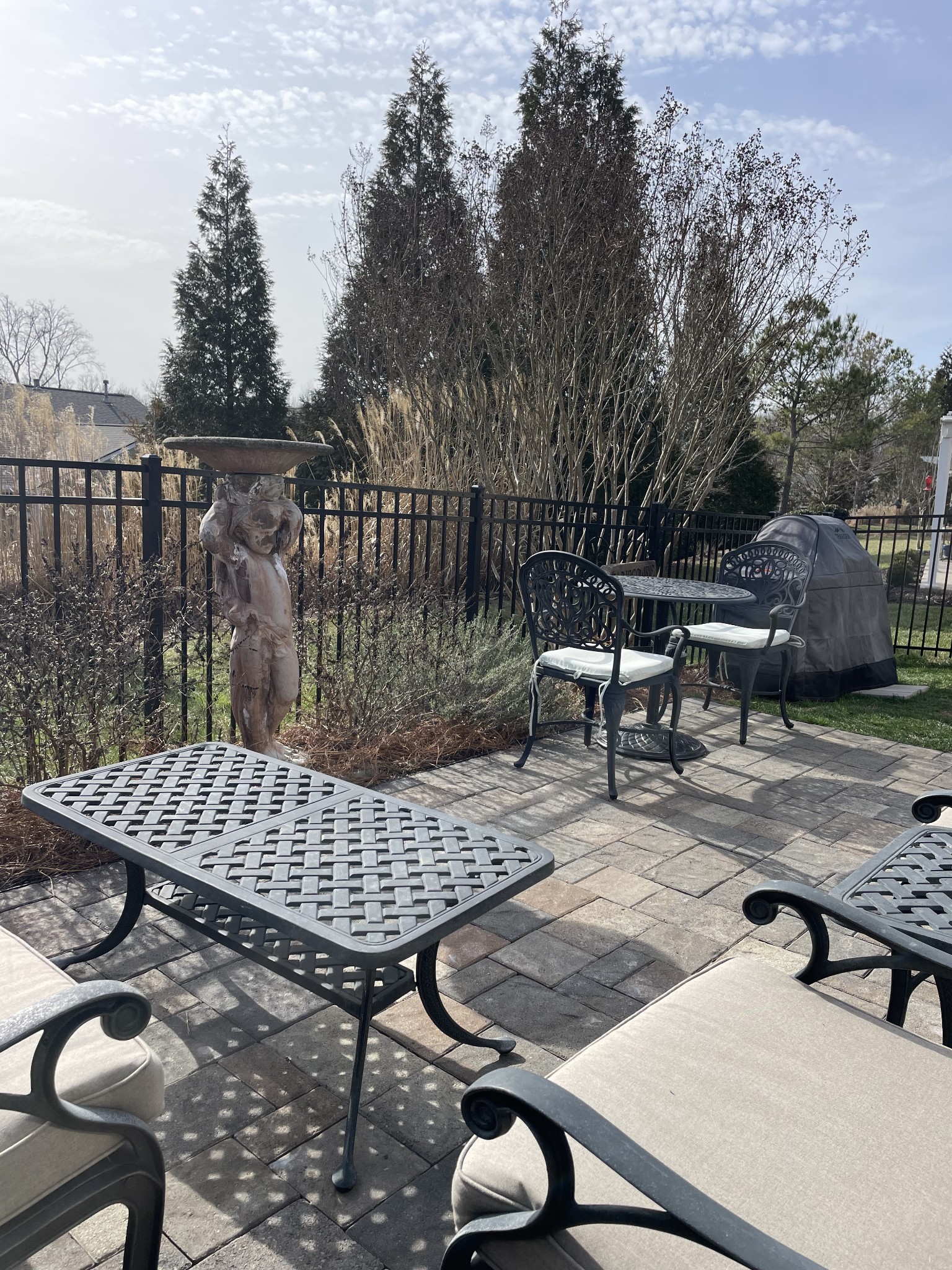 Paver patio with black metal seating, a stone birdbath, and a covered grill beside the garden fence.