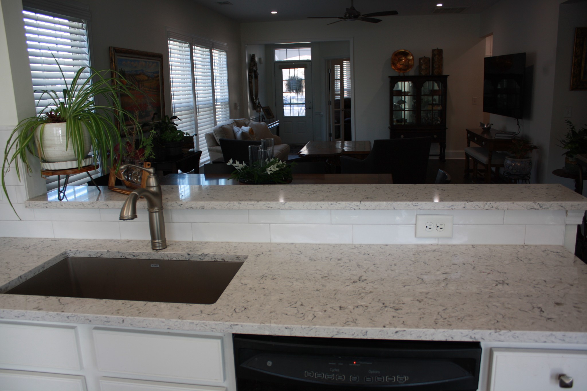 Kitchen sink and granite counter overlooking a furnished living room with shuttered windows and a wall-mounted television.