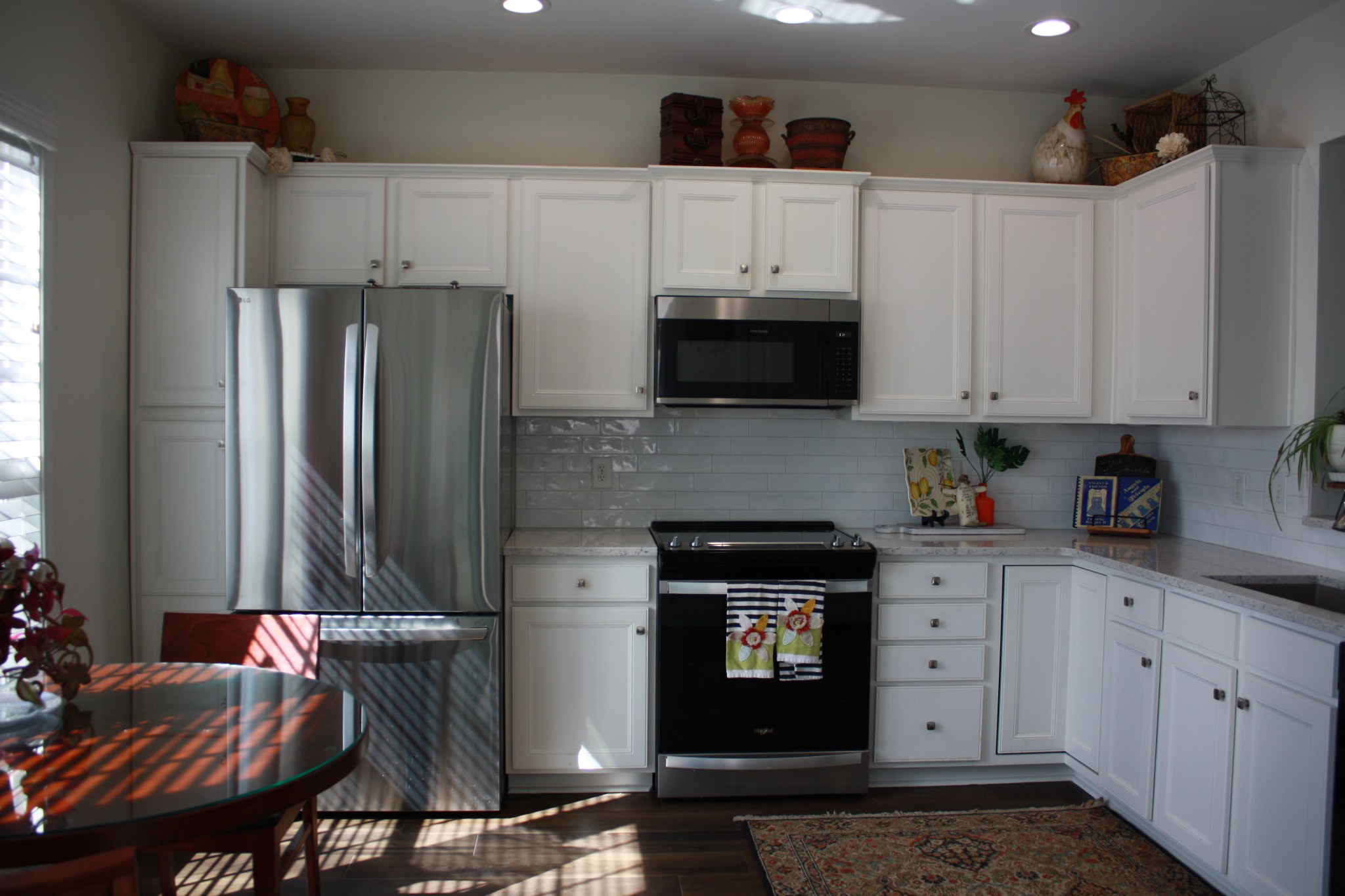 Kitchen with white cabinets, stainless appliances, gray tile backsplash, and sunlight casting striped shadows across a glass dining table.