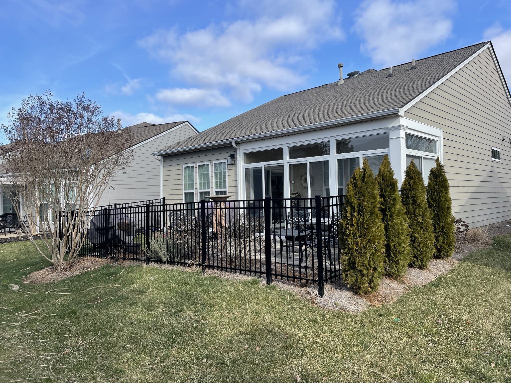 Rear of a gray house with a glass sunroom, fenced patio, outdoor chairs, and evergreen shrubs.