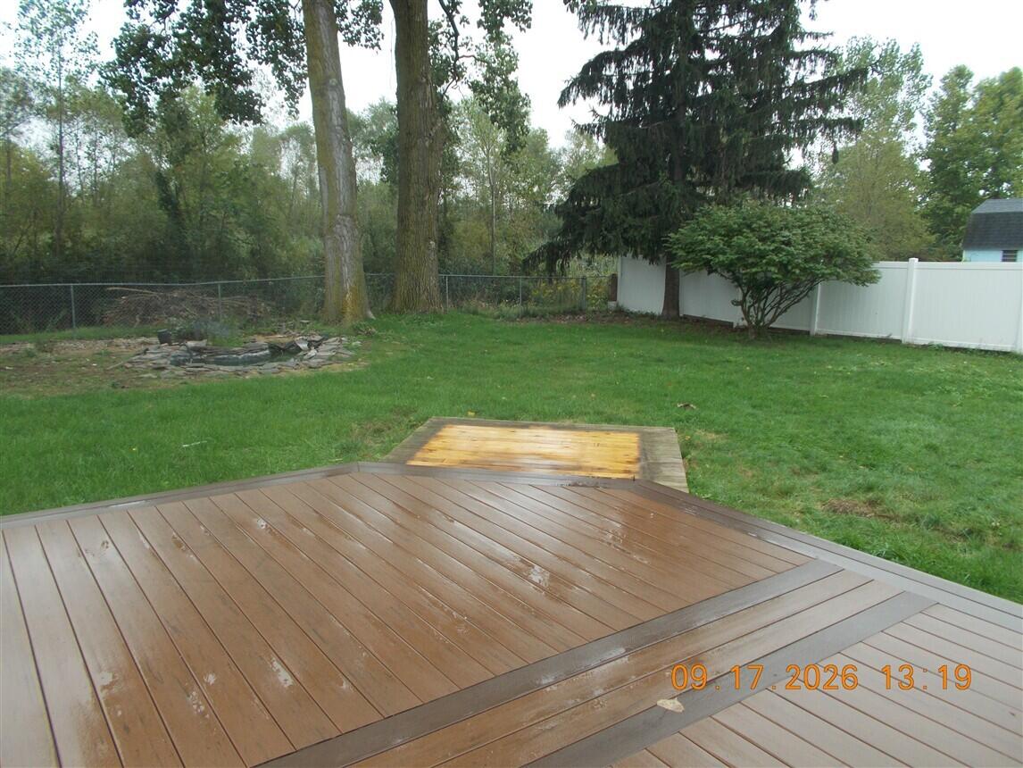 Wooden backyard deck overlooking a fenced lawn with mature trees and a stone fire pit.