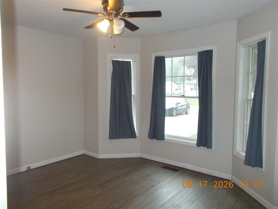 Empty room with dark wood flooring, three curtained windows, and a ceiling fan.