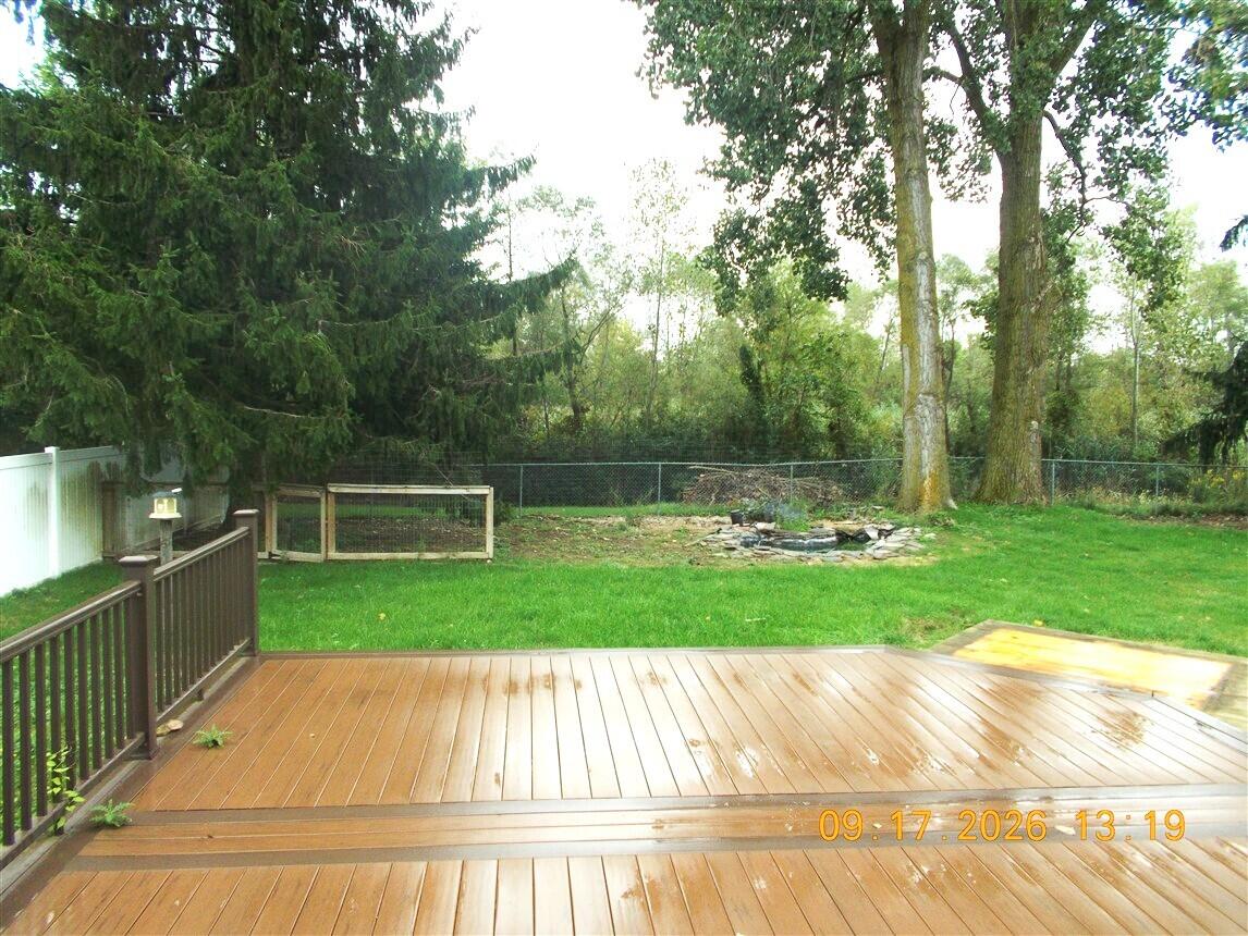 Backyard with a wooden deck, fenced lawn, stone fire pit, and mature trees.
