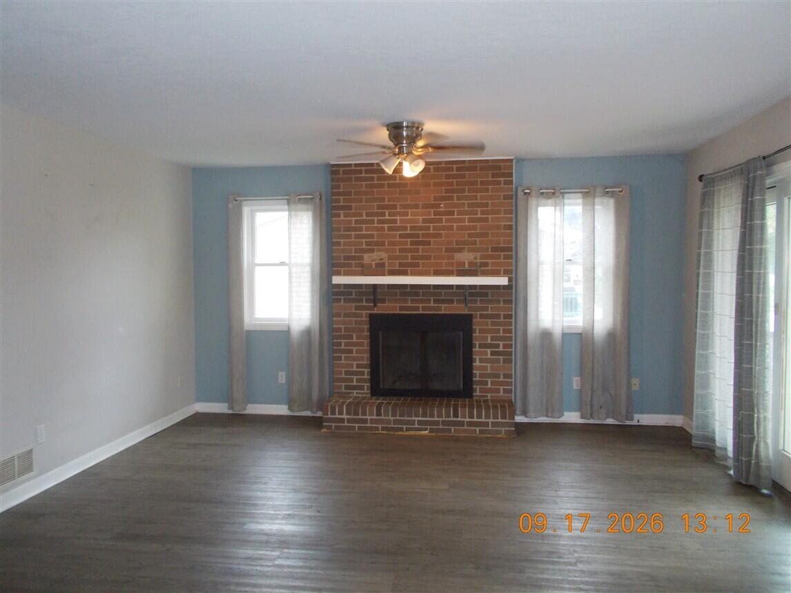 Empty living room with a brick fireplace, blue walls, dark flooring, and a ceiling fan.