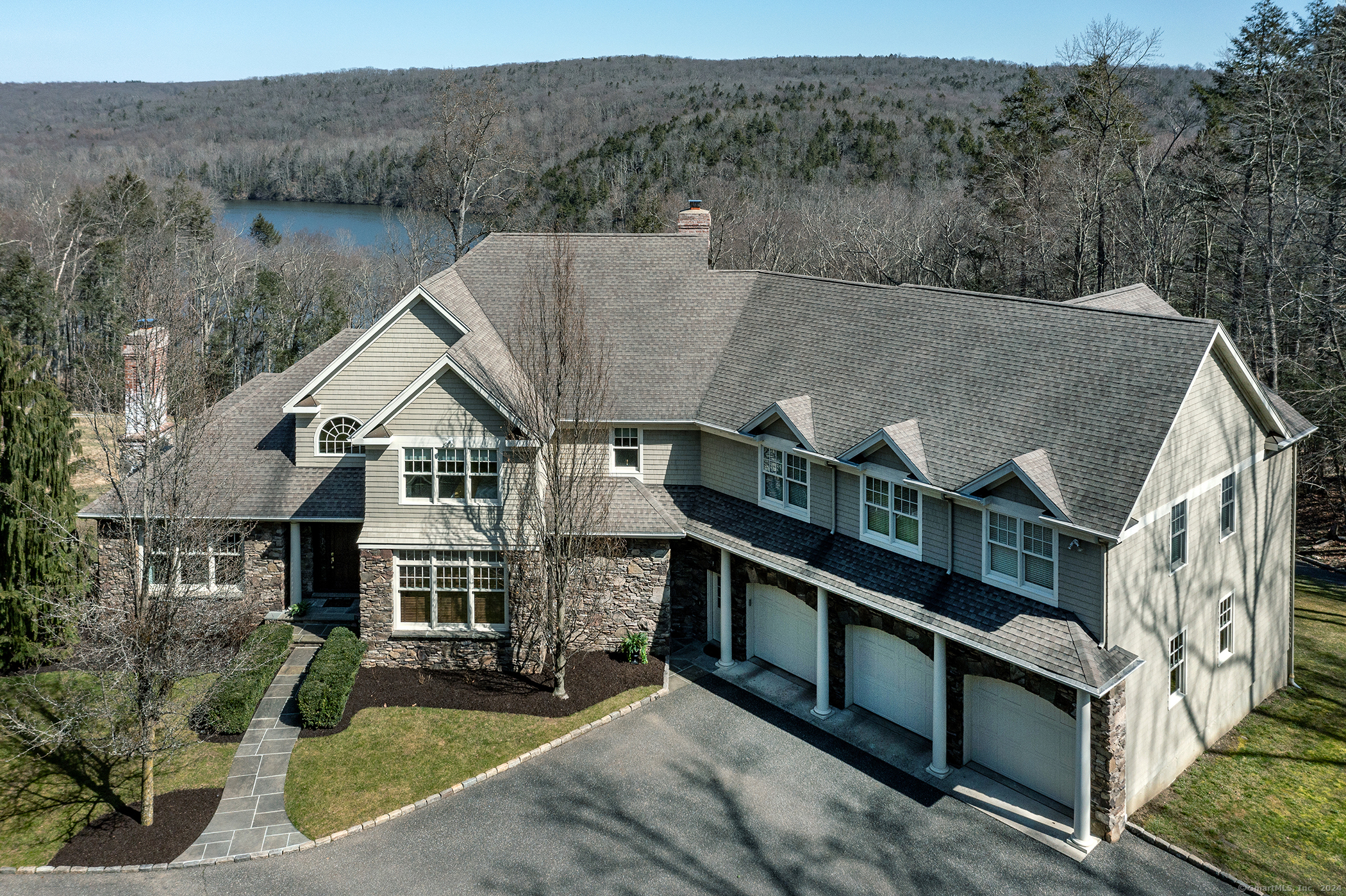Large gray-shingled home with stone accents and a three-car garage, overlooking a lake.
