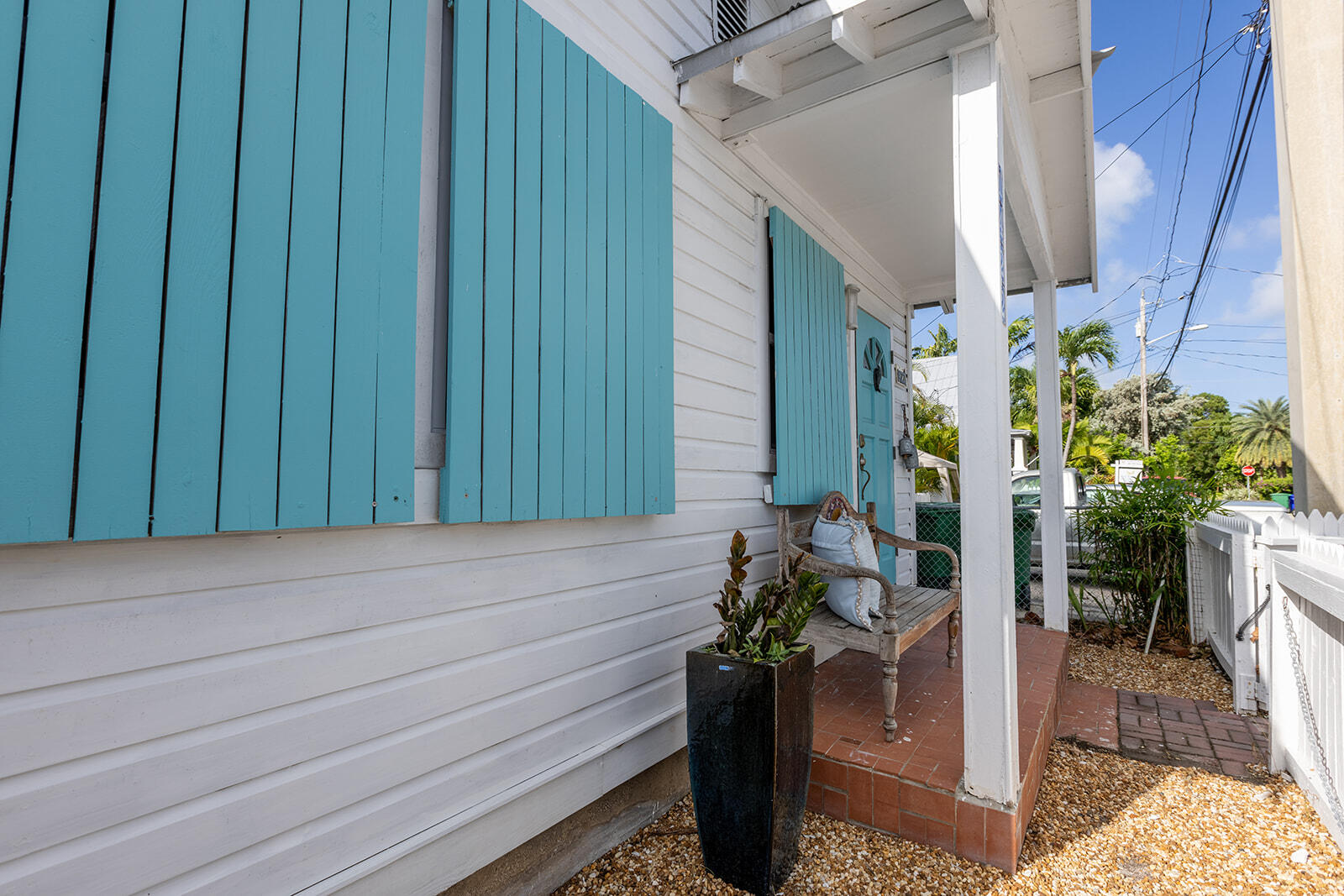 White house porch with turquoise shutters, a wooden bench, and a white picket fence.