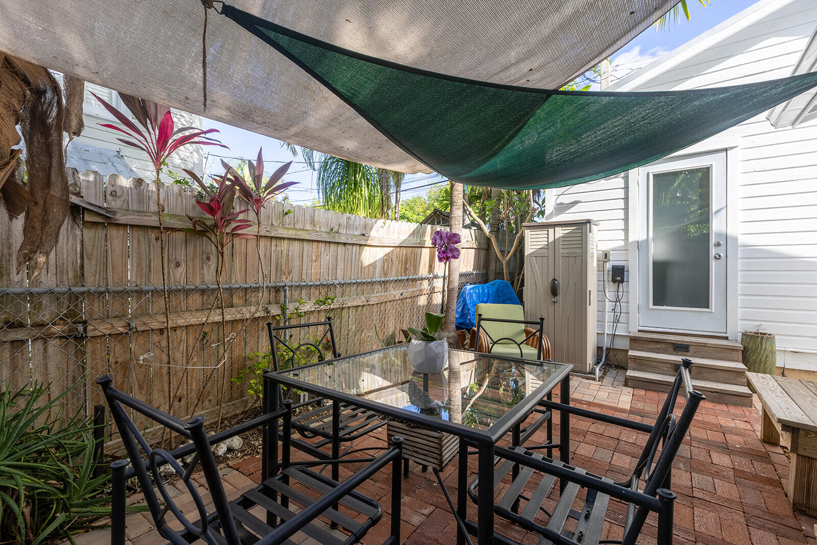 Brick patio with outdoor dining set beneath shade sails beside a white house.