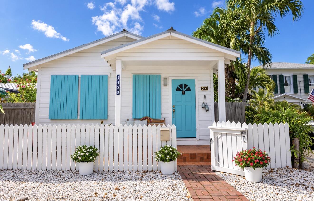 White cottage with turquoise shutters and door behind a picket fence and tropical landscaping.
