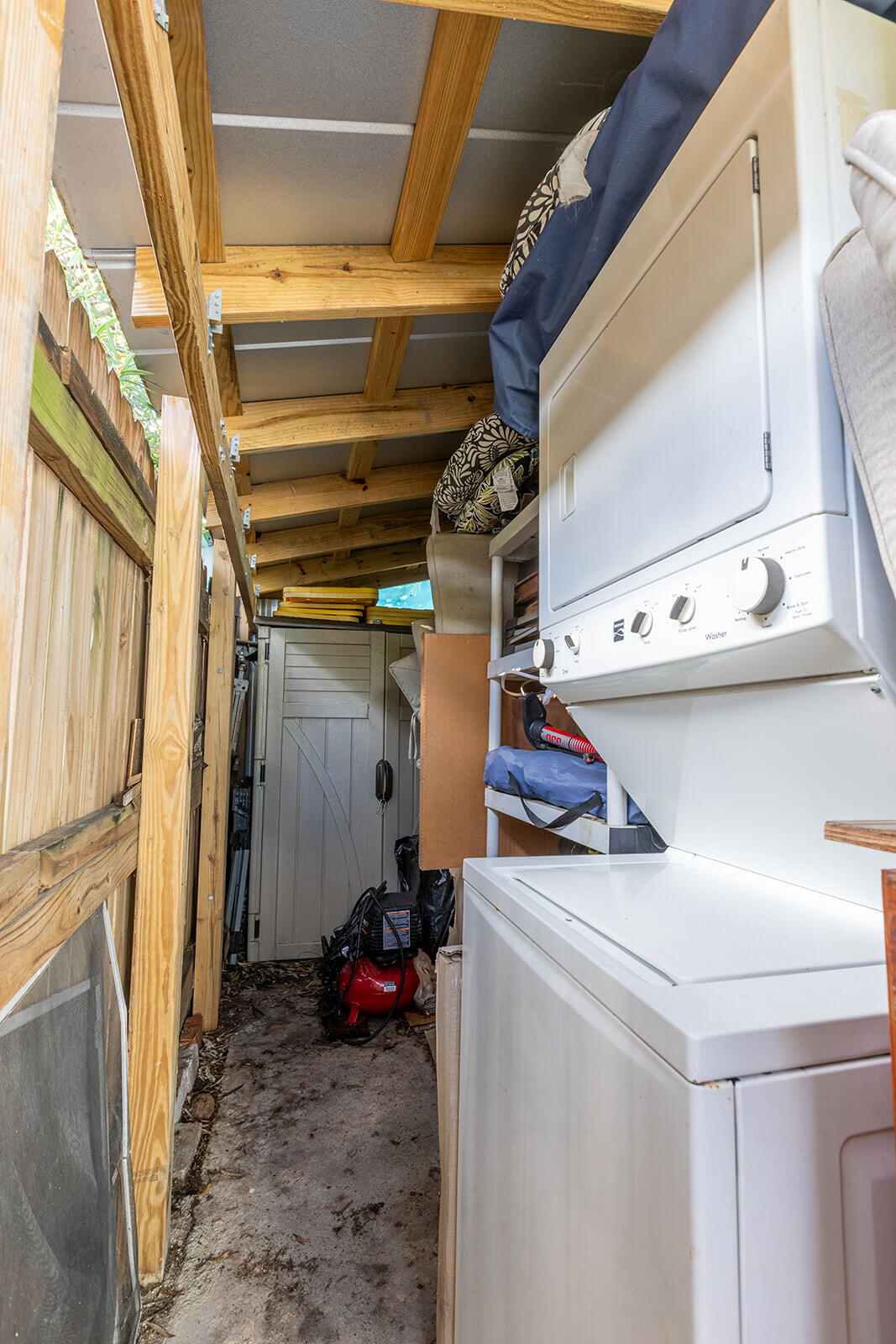 Narrow laundry area with stacked washer and dryer beside exposed wooden framing and overhead storage.