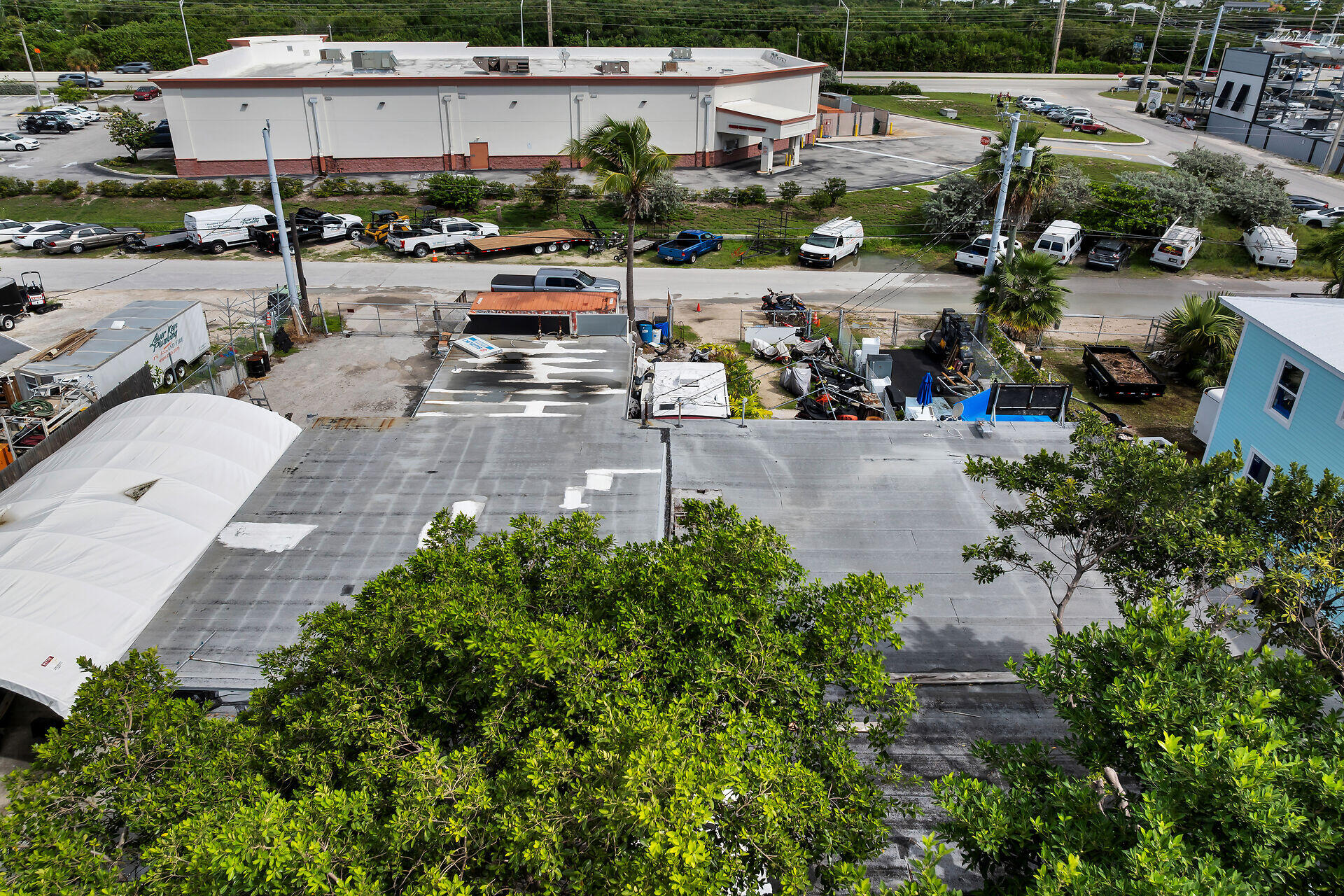 Aerial view of a flat-roofed property behind tropical trees, with a fenced work area and marina nearby.