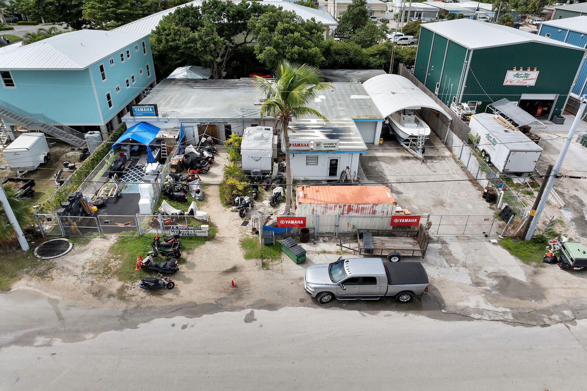 Aerial view of a marine repair yard with covered boats, scooters, trailers, and a palm tree.