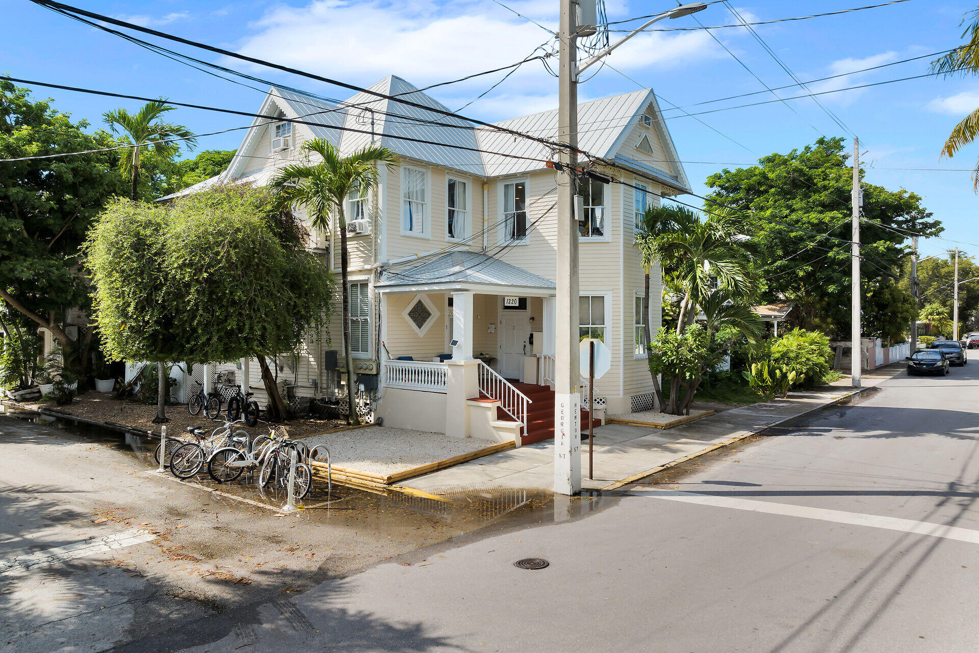 Two-story cream house with a silver metal roof, covered porch, tropical landscaping, and bicycles along the sidewalk.