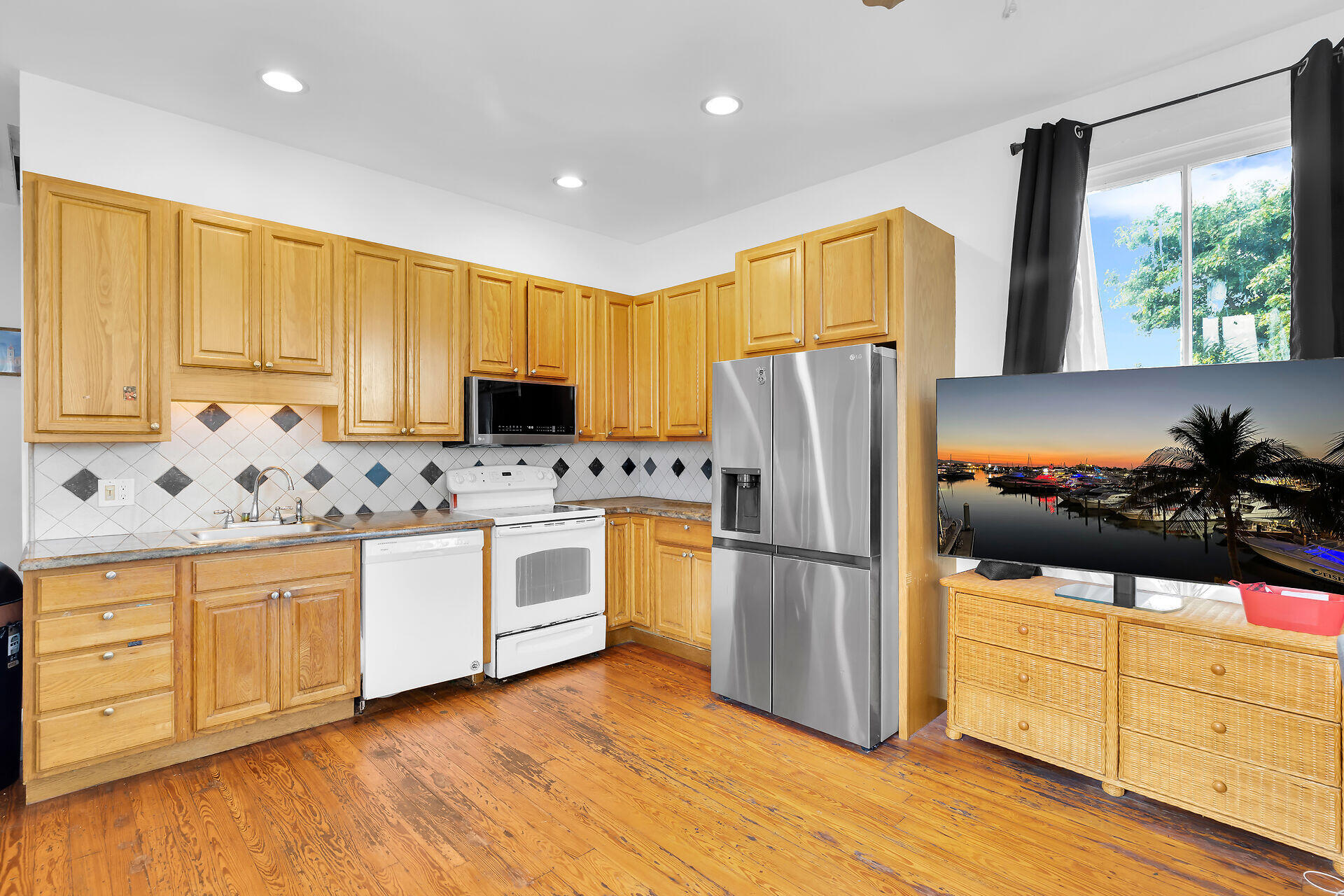 Kitchen with honey-colored cabinets, stainless steel refrigerator, wood floors, and a television displaying a marina at sunset.