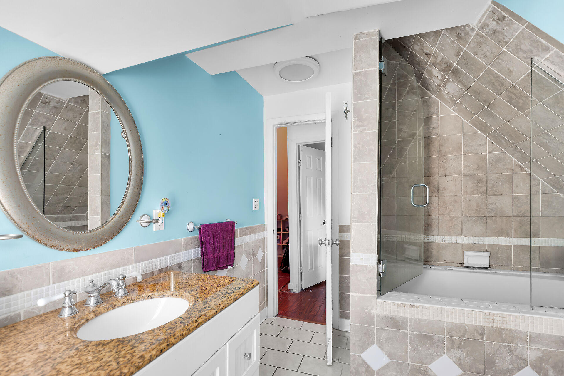 Bathroom with a glass-enclosed tub and shower, turquoise wall, and granite vanity beneath an oval mirror.