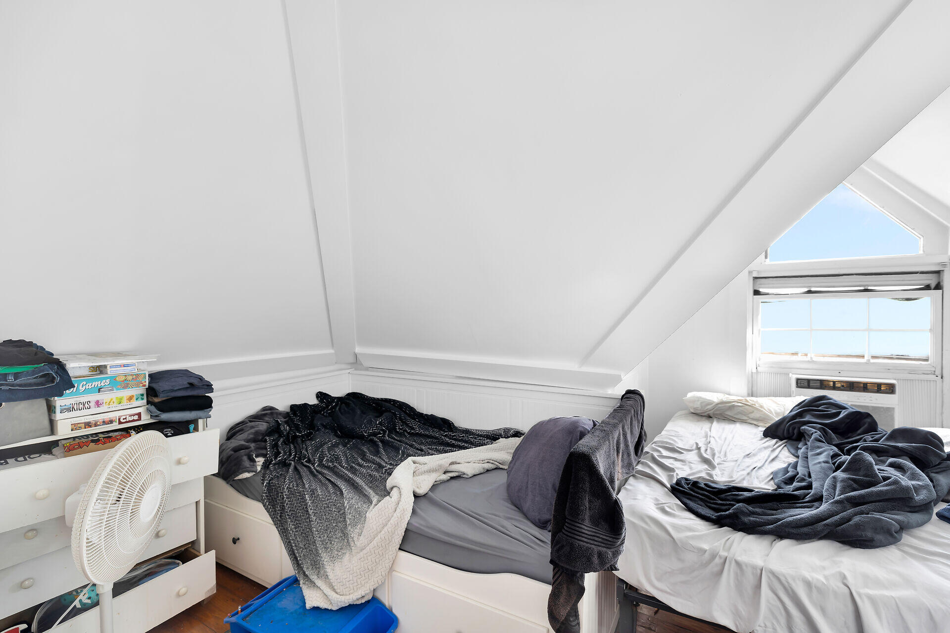 Bedroom with two unmade beds beneath a sloped ceiling and a bright window.