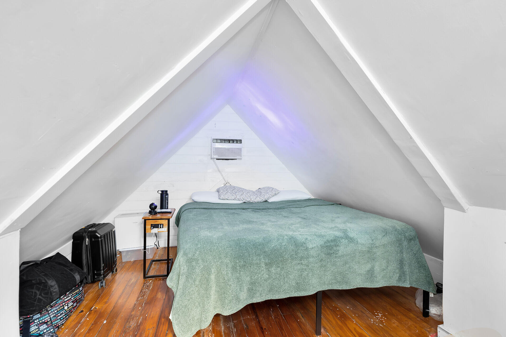 Attic bedroom with a green-covered bed, steep white ceiling, hardwood floors, and luggage.
