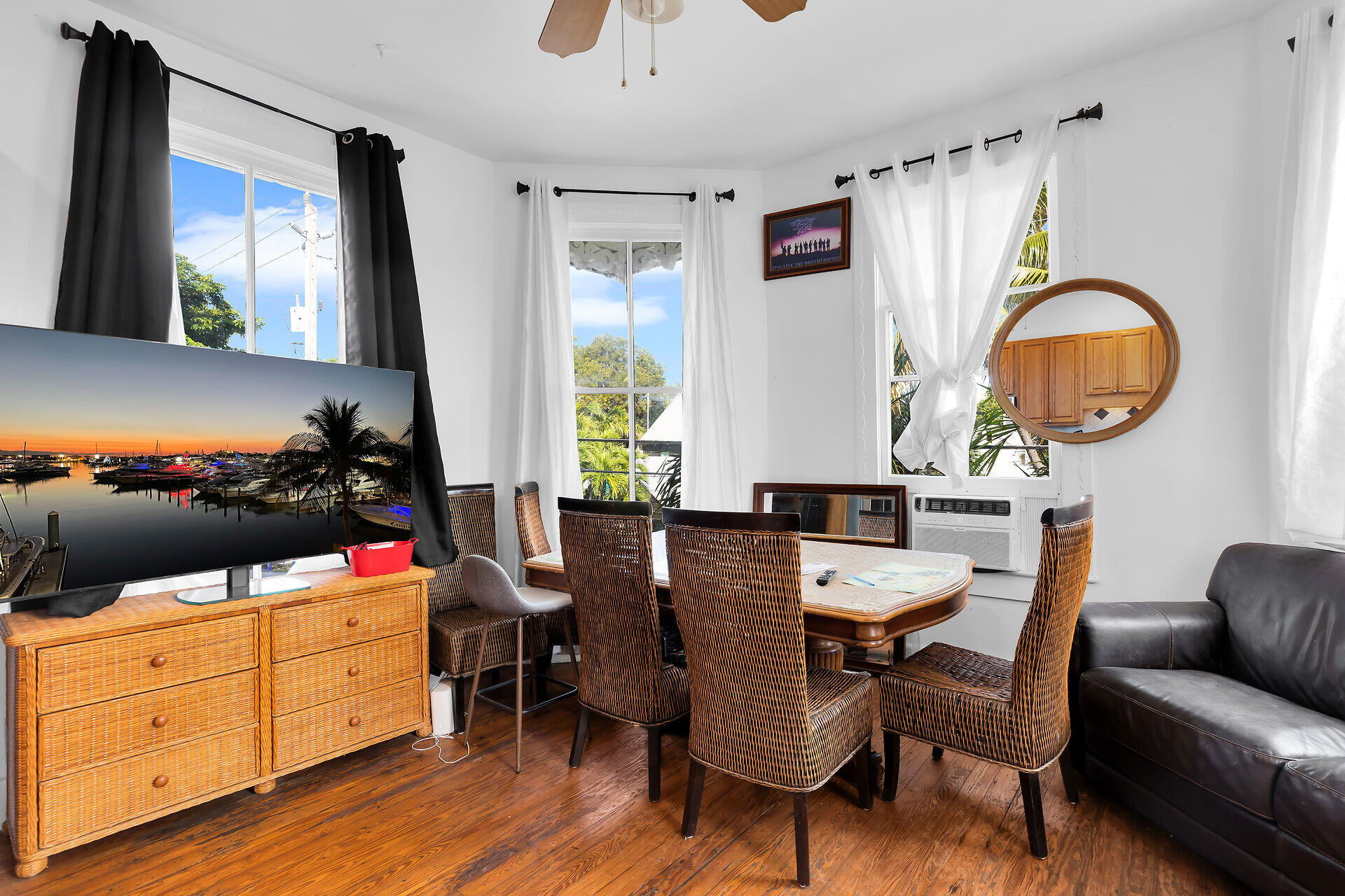Living and dining area with wicker furniture, polished wood floors, and tall curtained windows.