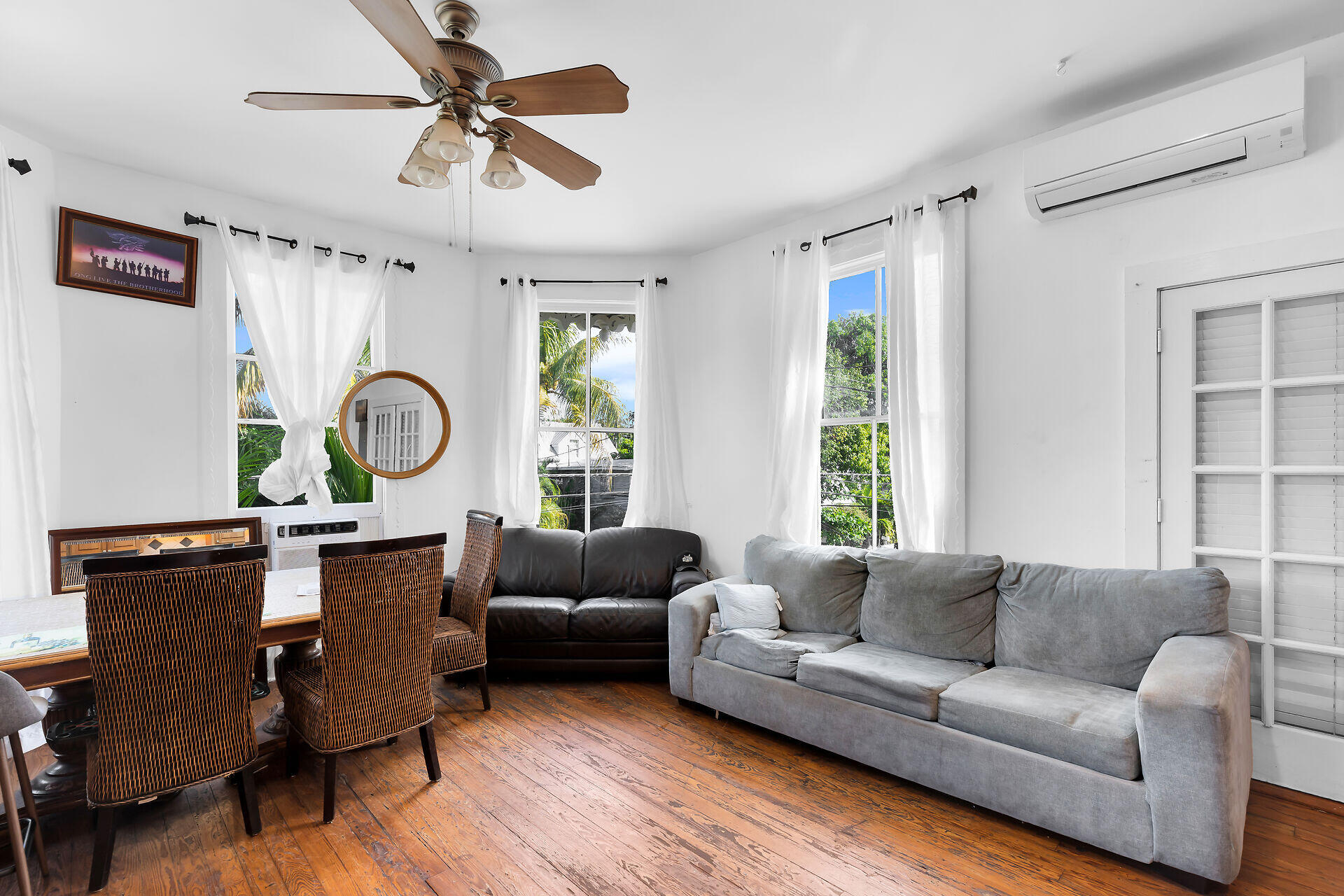 Living and dining room with wicker chairs, gray sofas, and windows overlooking tropical greenery.