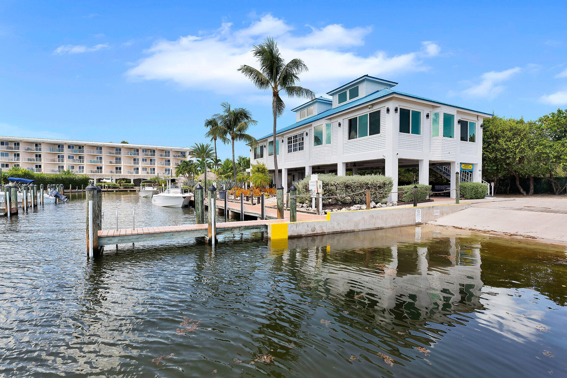 Waterfront building with a blue roof, marina docks, boats, and palm trees.