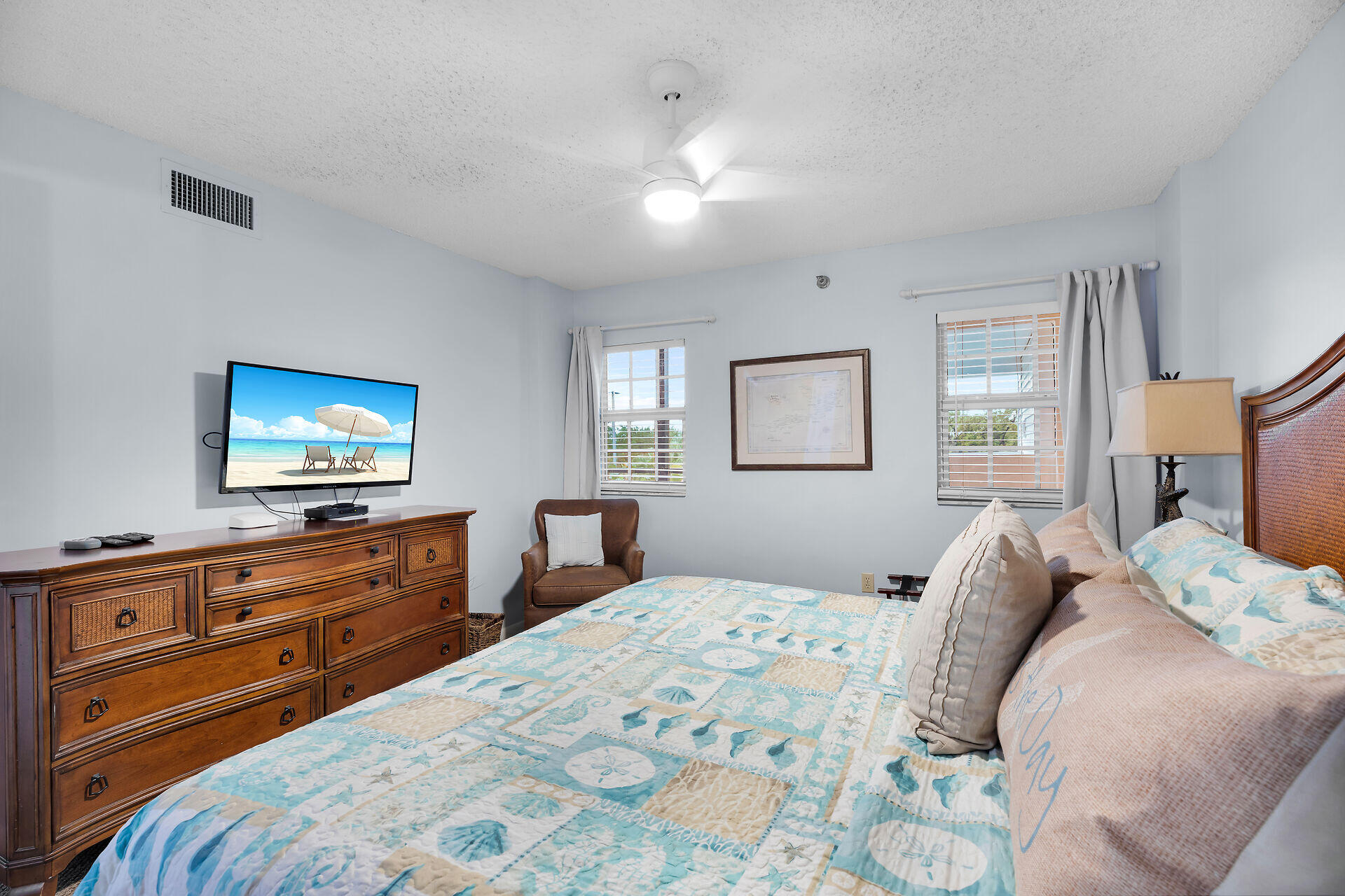 Furnished bedroom with a seashell-patterned quilt, dark wood dresser, television, and two curtained windows.