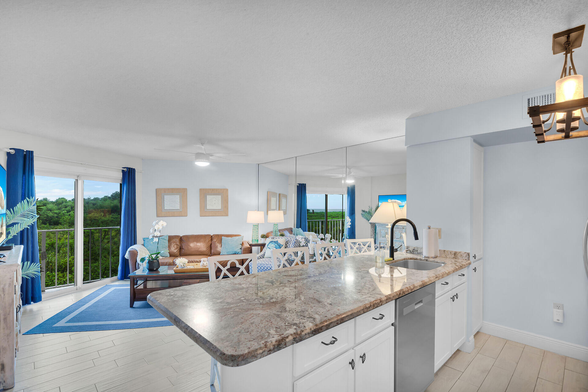 Open kitchen with a granite island and furnished living area beside windows overlooking greenery.