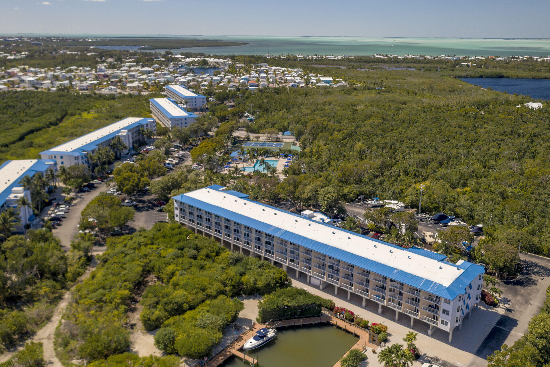 Aerial view of waterfront condominiums, a pool and tennis courts beside mangroves and turquoise water.