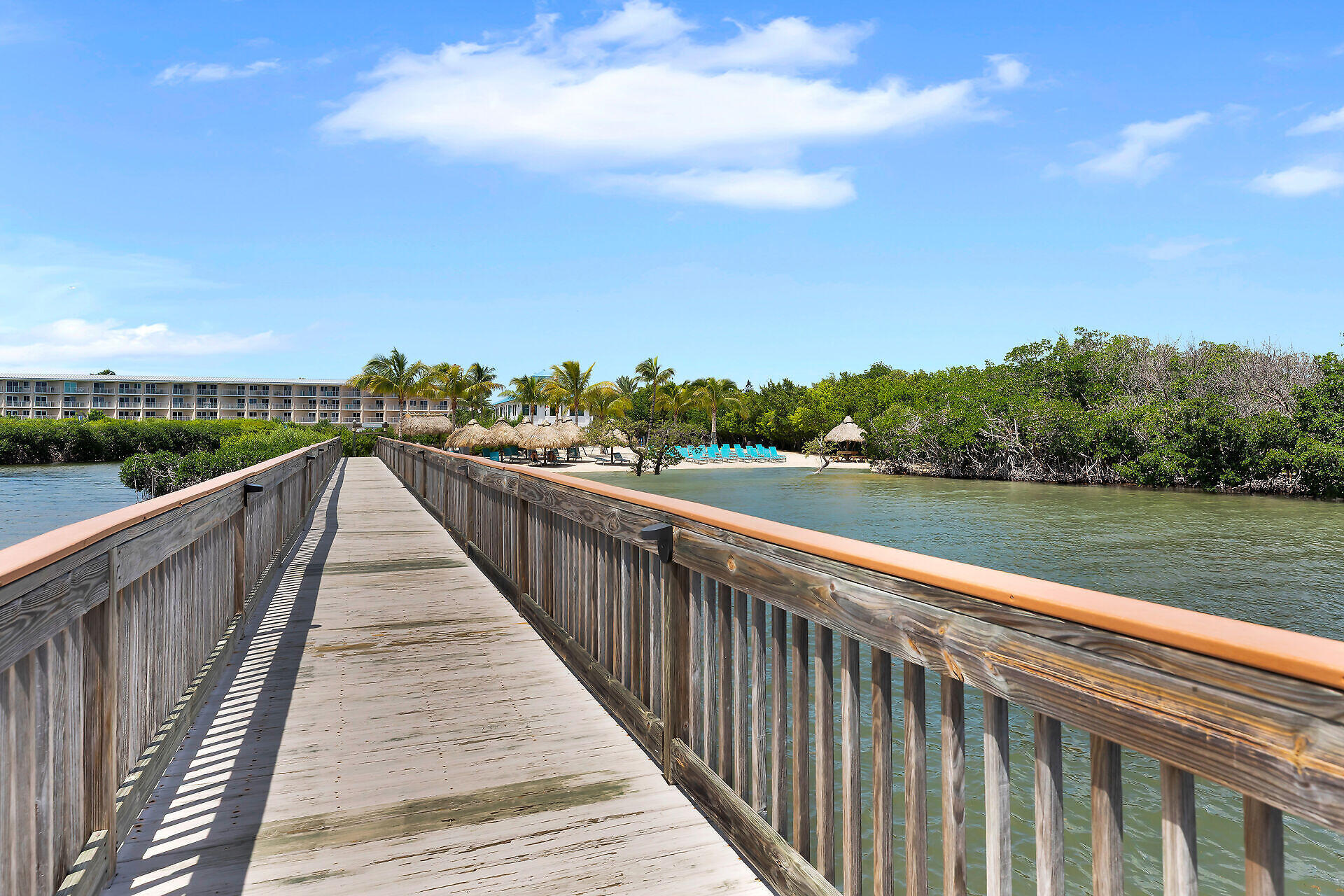 Wooden boardwalk over a channel leads to a resort pool with turquoise chairs and thatched umbrellas.