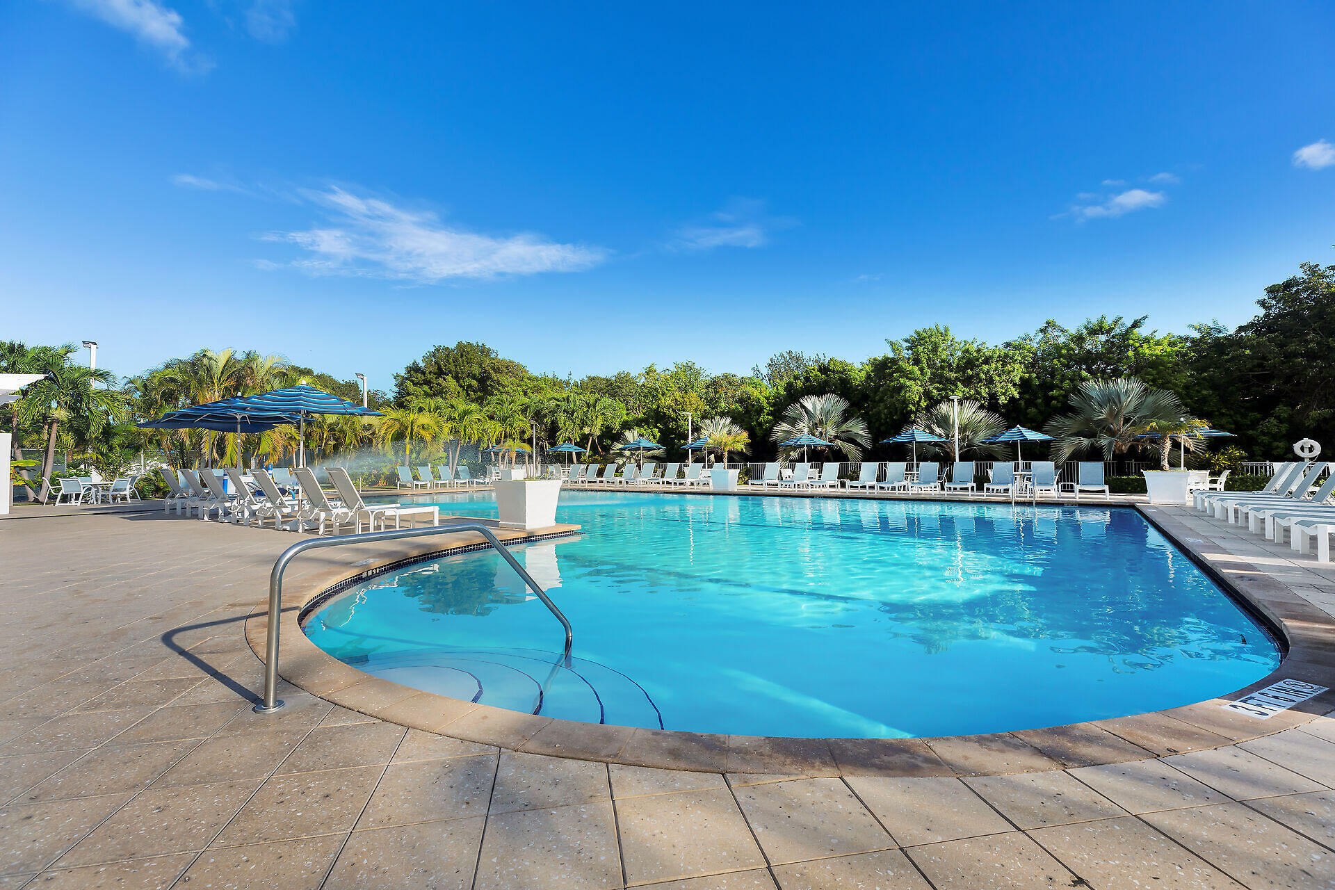 Outdoor swimming pool with white lounge chairs, striped umbrellas, and tropical landscaping.