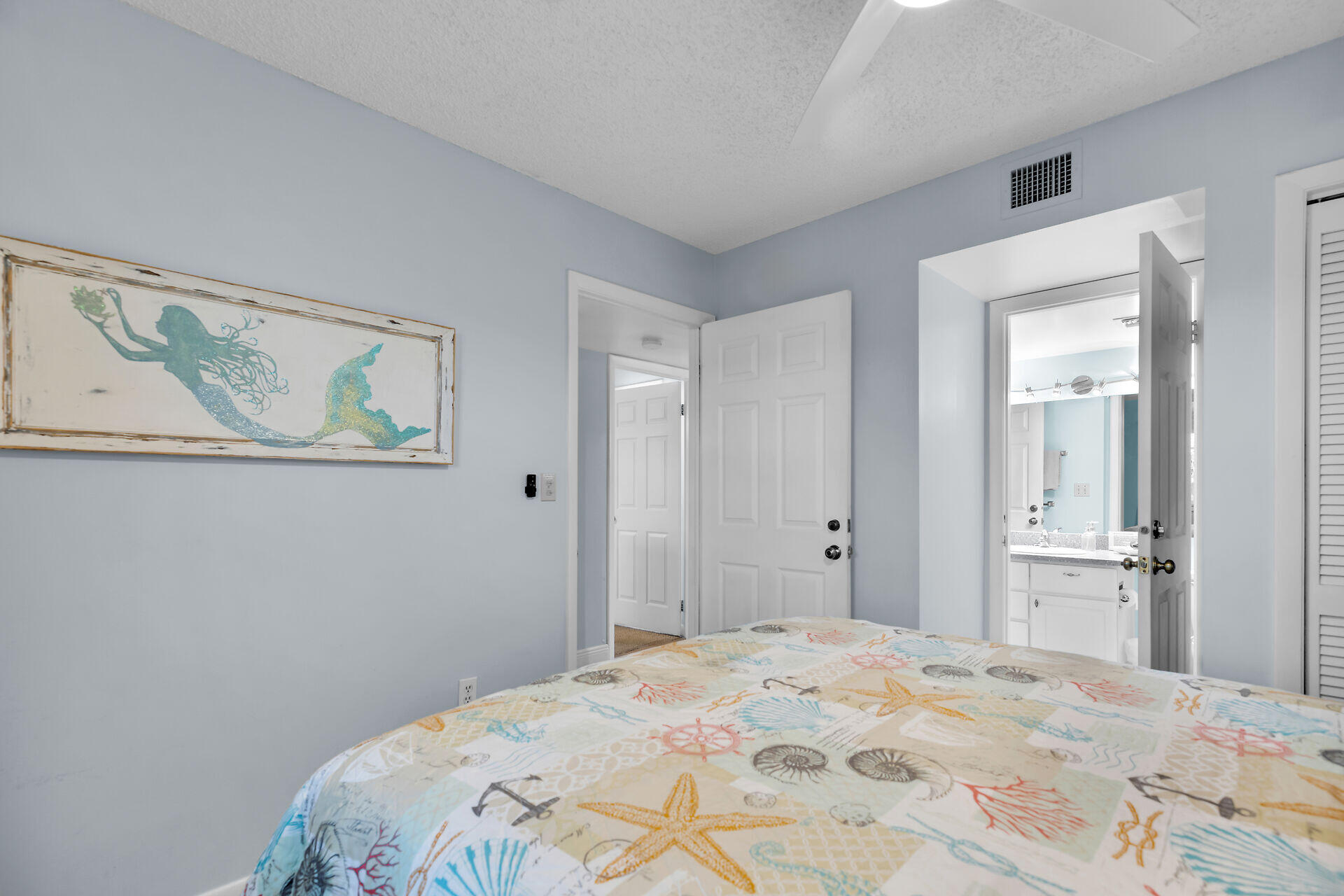 Furnished bedroom with marine-themed bedding and framed mermaid artwork, opening to a bathroom and hallway.