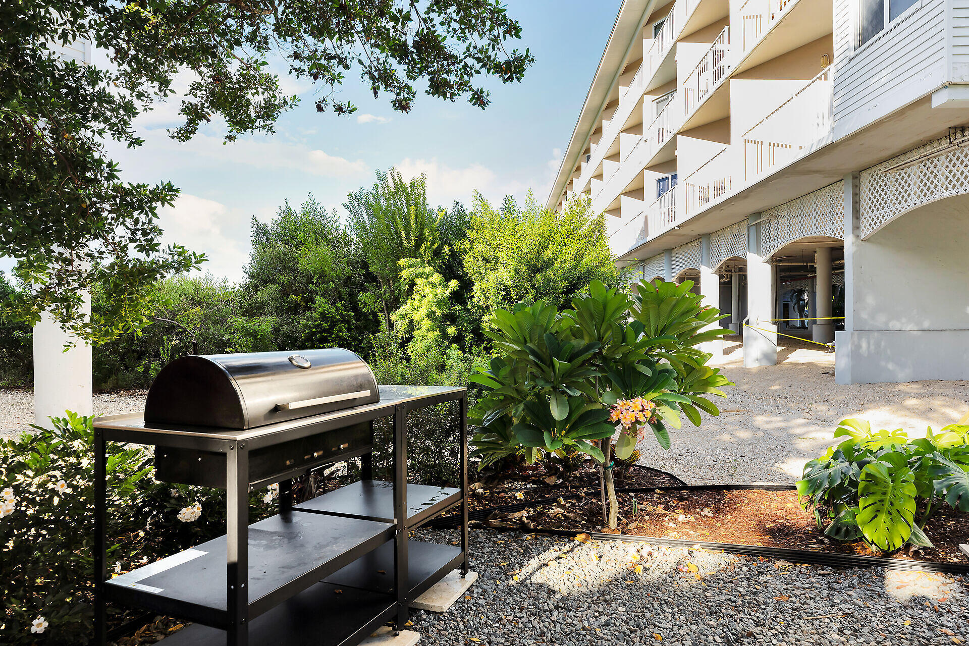 Condominium exterior with white balconies, tropical landscaping, and an outdoor stainless-steel grill.