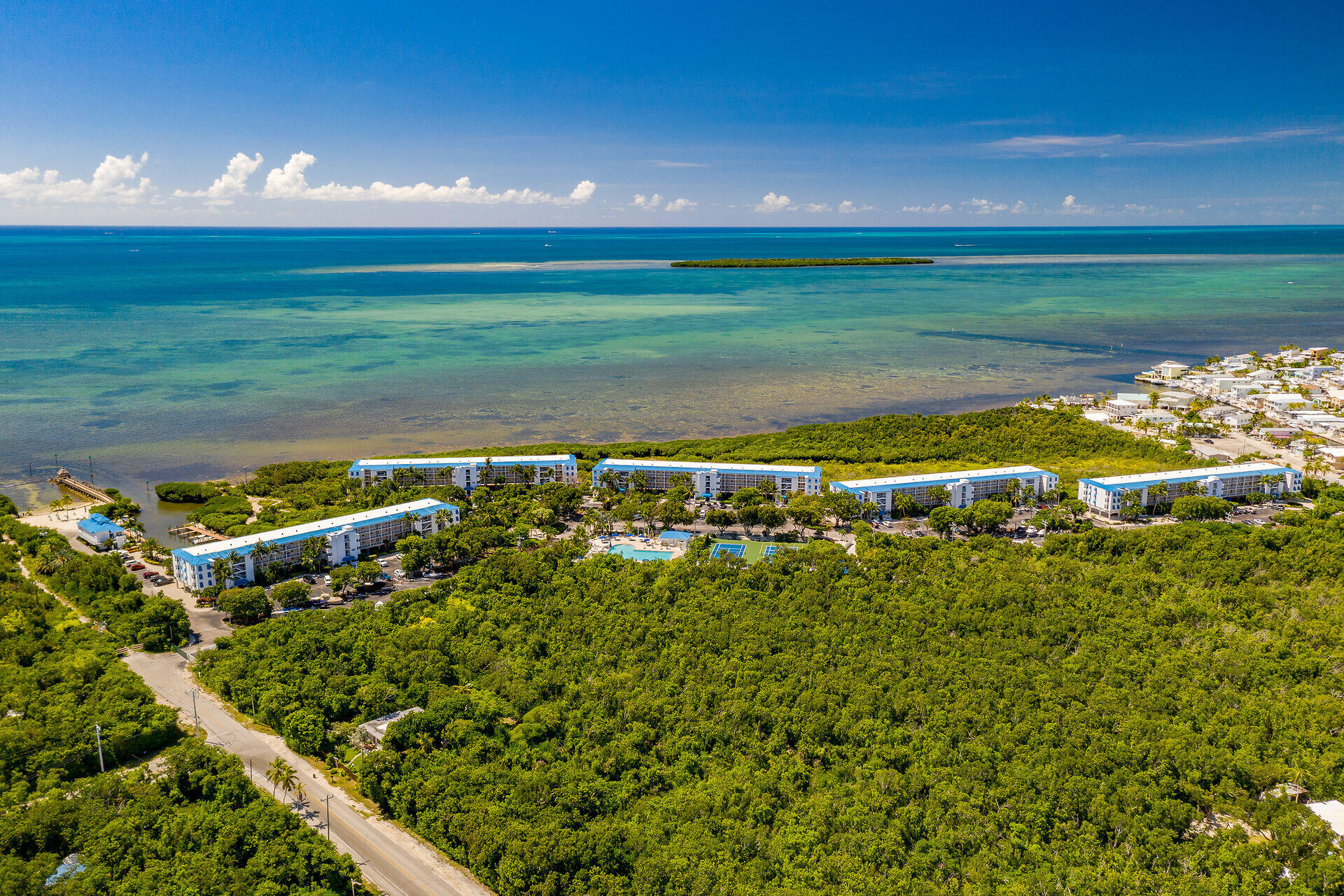 Aerial view of waterfront condominiums beside turquoise ocean, mangroves, a pool, and tennis courts.