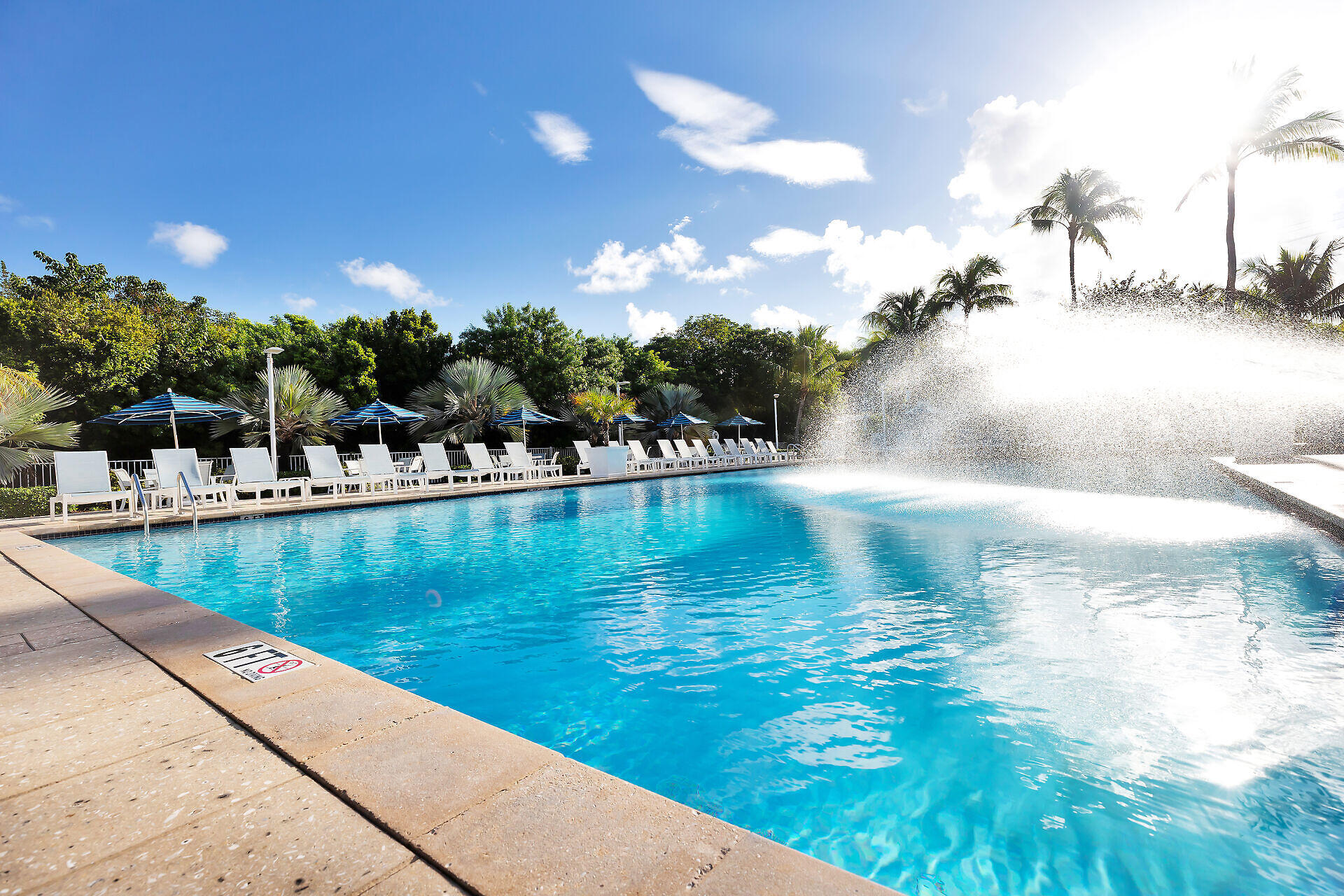 Outdoor pool with a broad fountain, white loungers, blue umbrellas, and palm trees.
