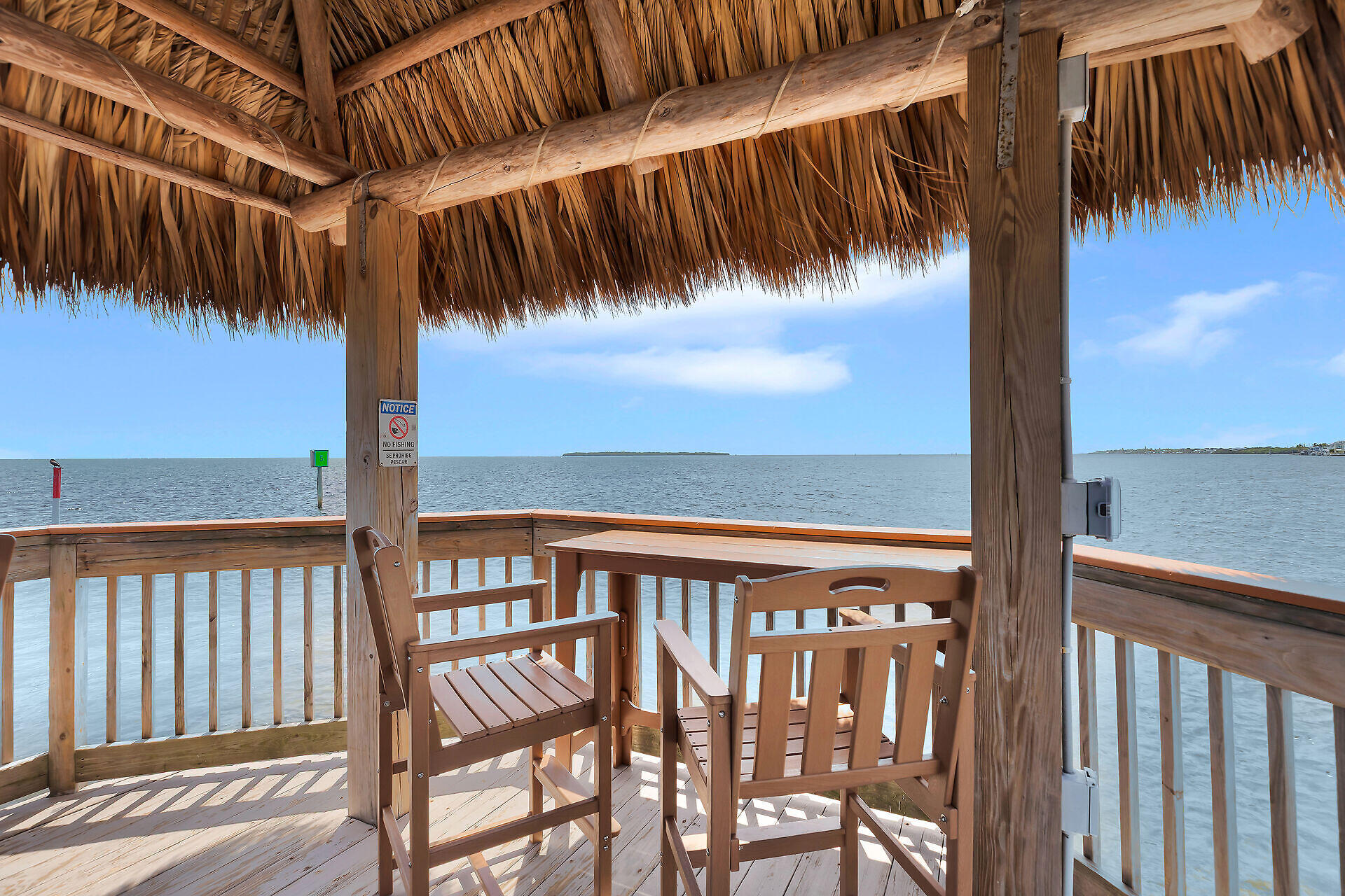 Waterfront deck with wooden seating beneath a thatched roof, overlooking calm water.
