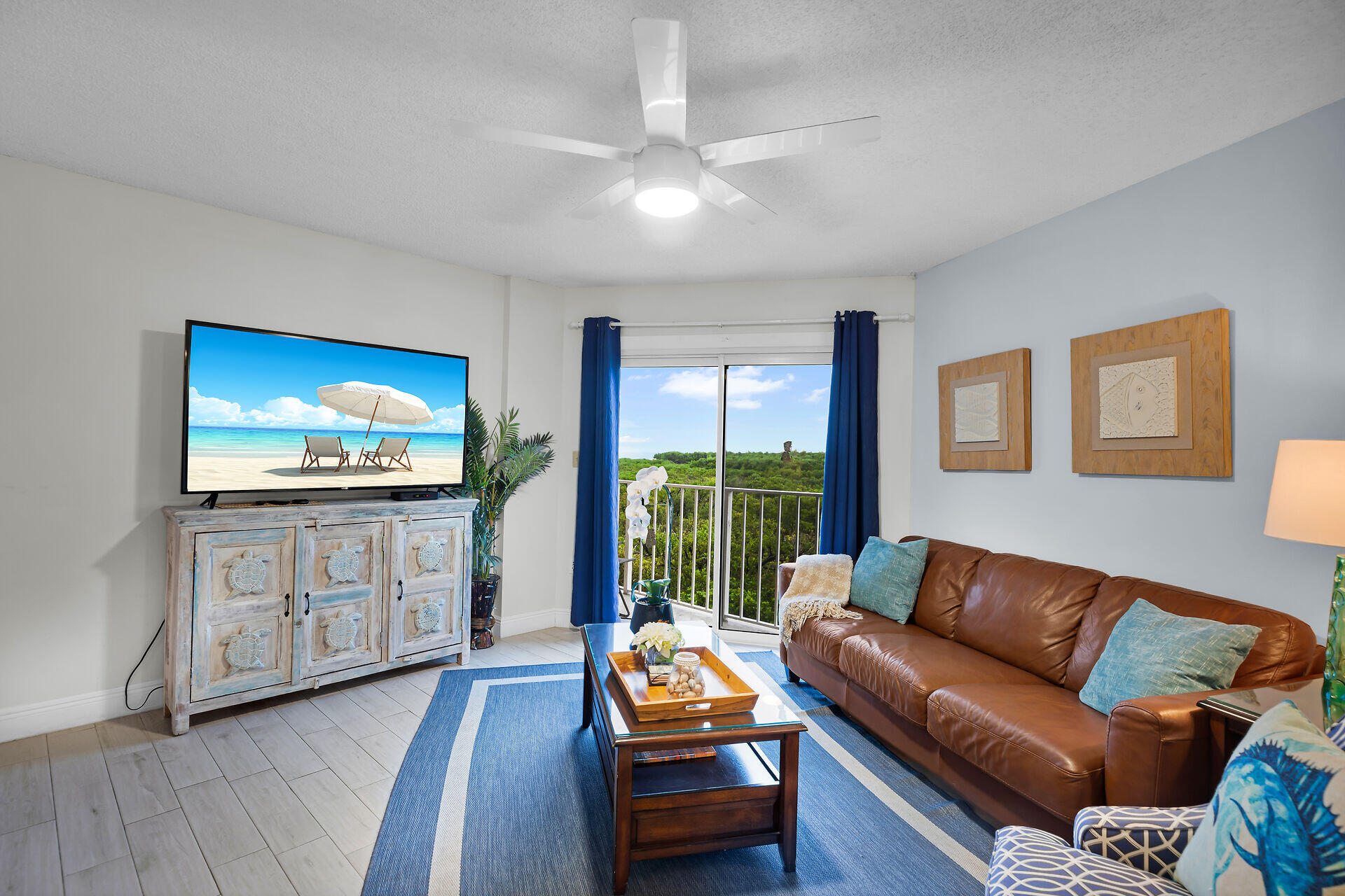 Furnished living room with a brown leather sectional and balcony doors overlooking green treetops.