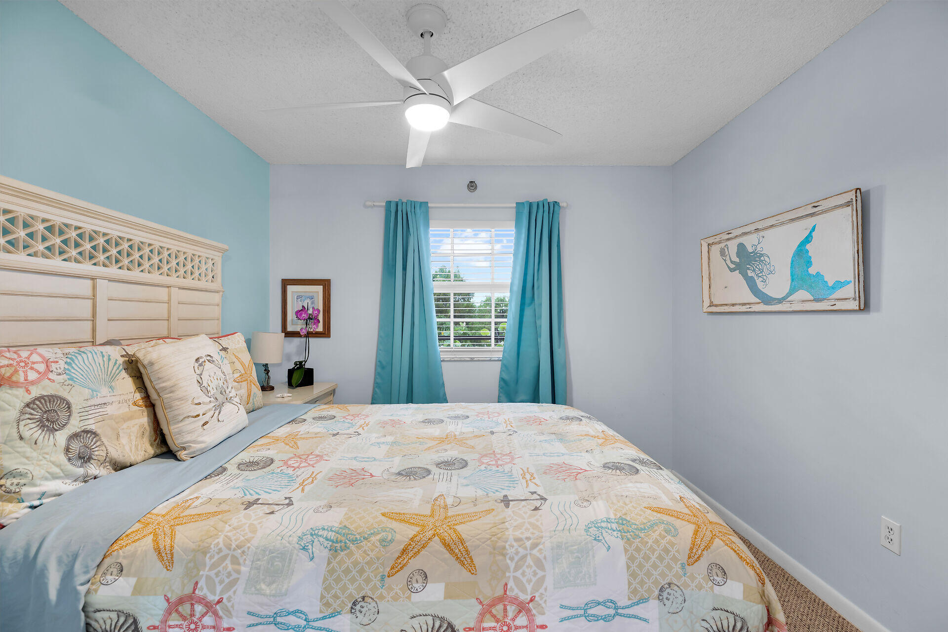 Bedroom with coastal-patterned bedding, turquoise curtains, a white ceiling fan, and mermaid wall art.