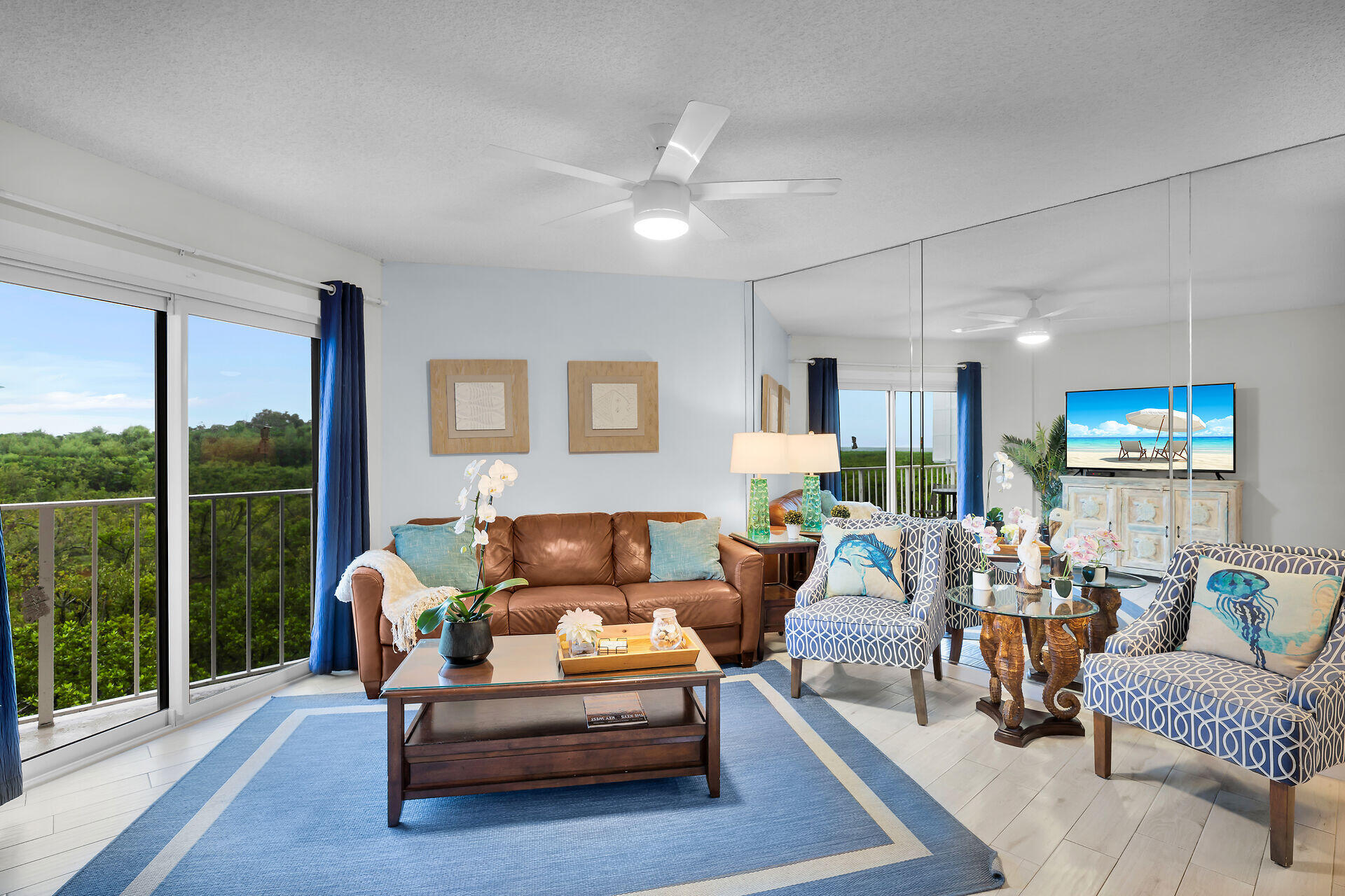 Furnished living room with a brown leather sofa, blue rug, mirrored wall, and sliding doors overlooking greenery.