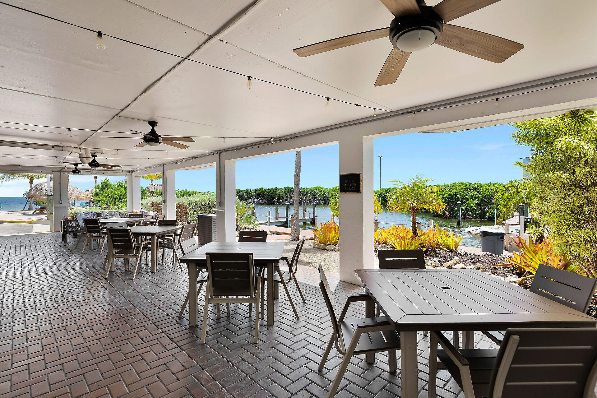 Covered waterfront patio with dining tables, ceiling fans, tropical landscaping, and a dock