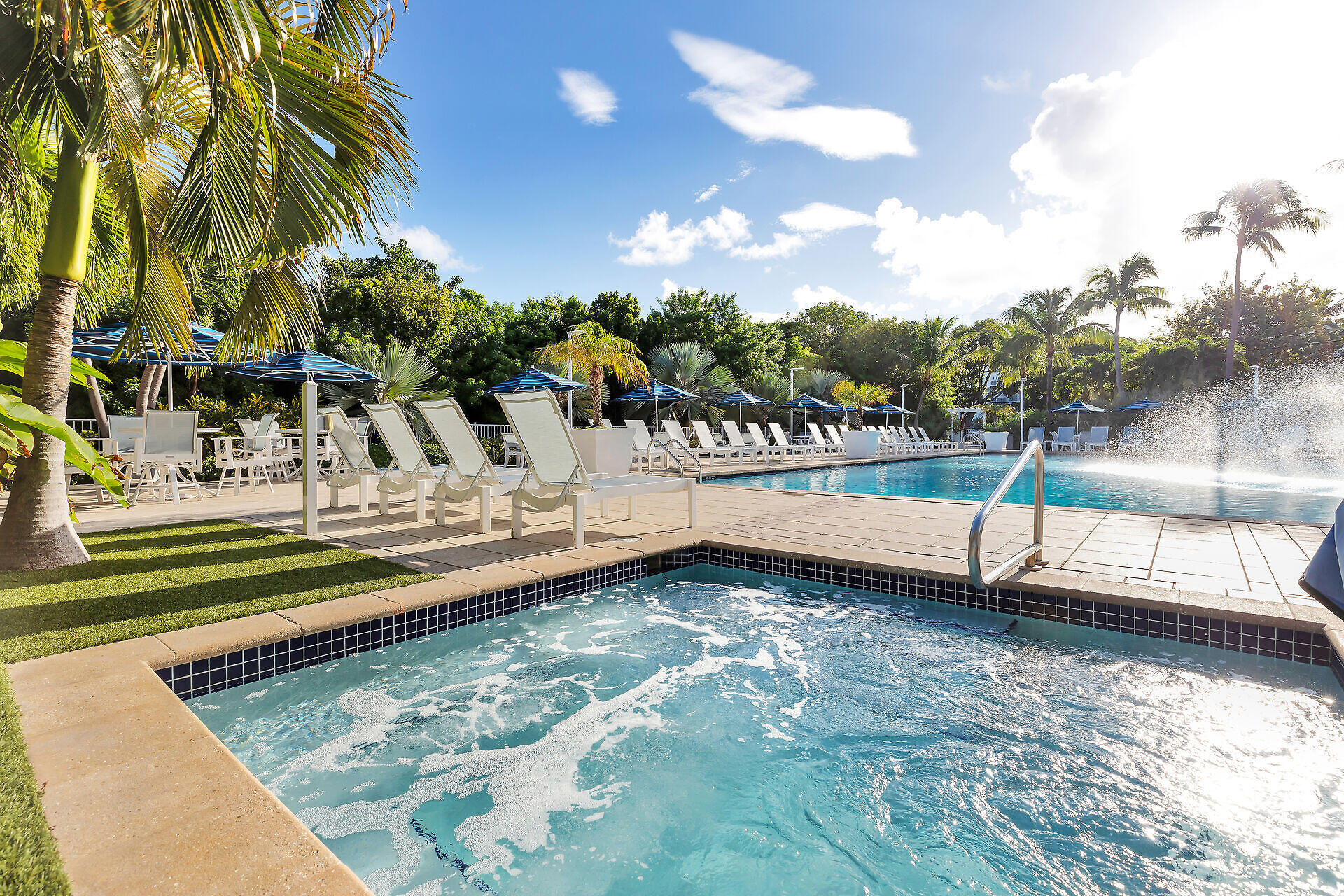 Outdoor swimming pool and bubbling spa beside lounge chairs, blue umbrellas, and palm trees.