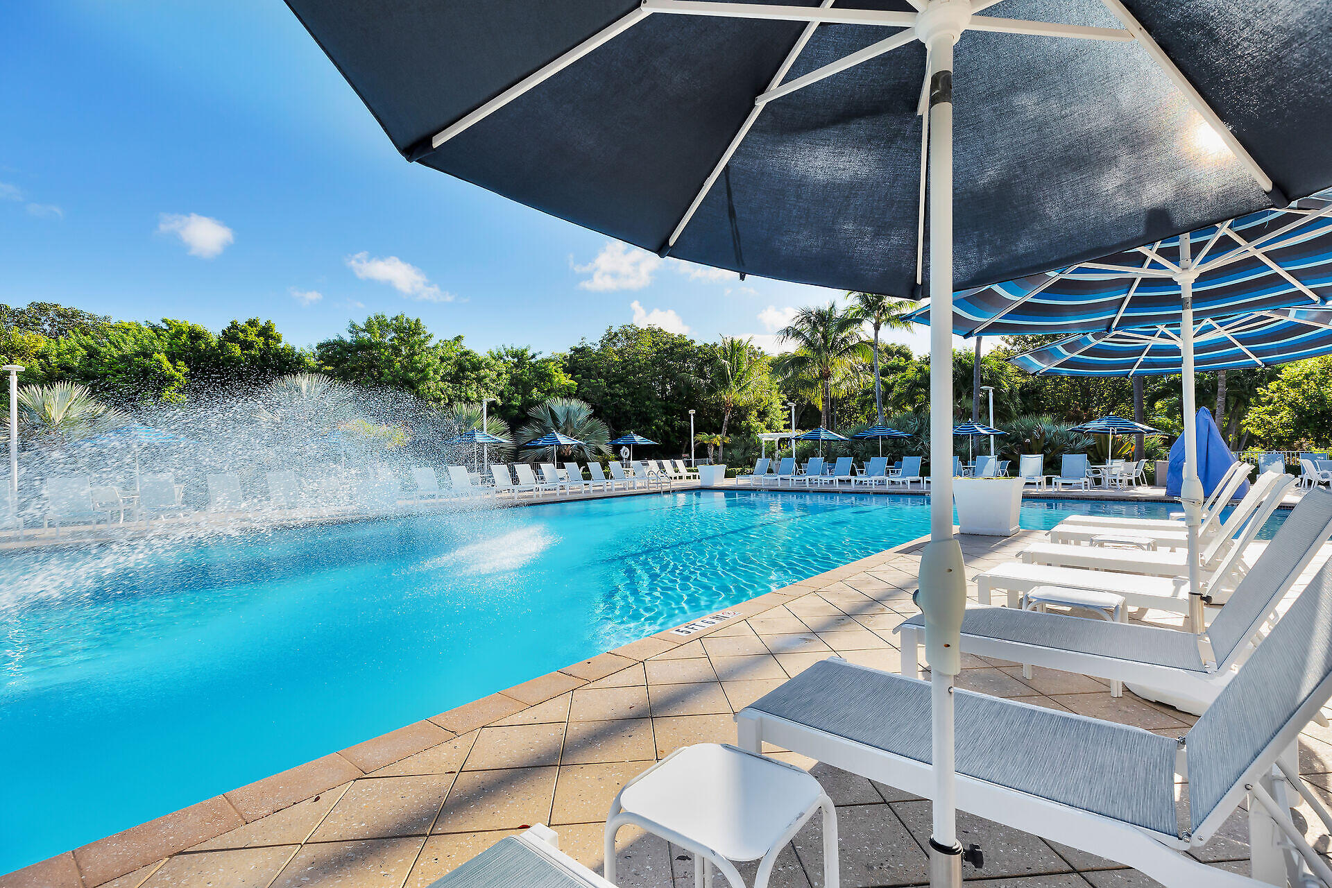 Outdoor pool with a spraying fountain, blue umbrellas, and rows of white lounge chairs.