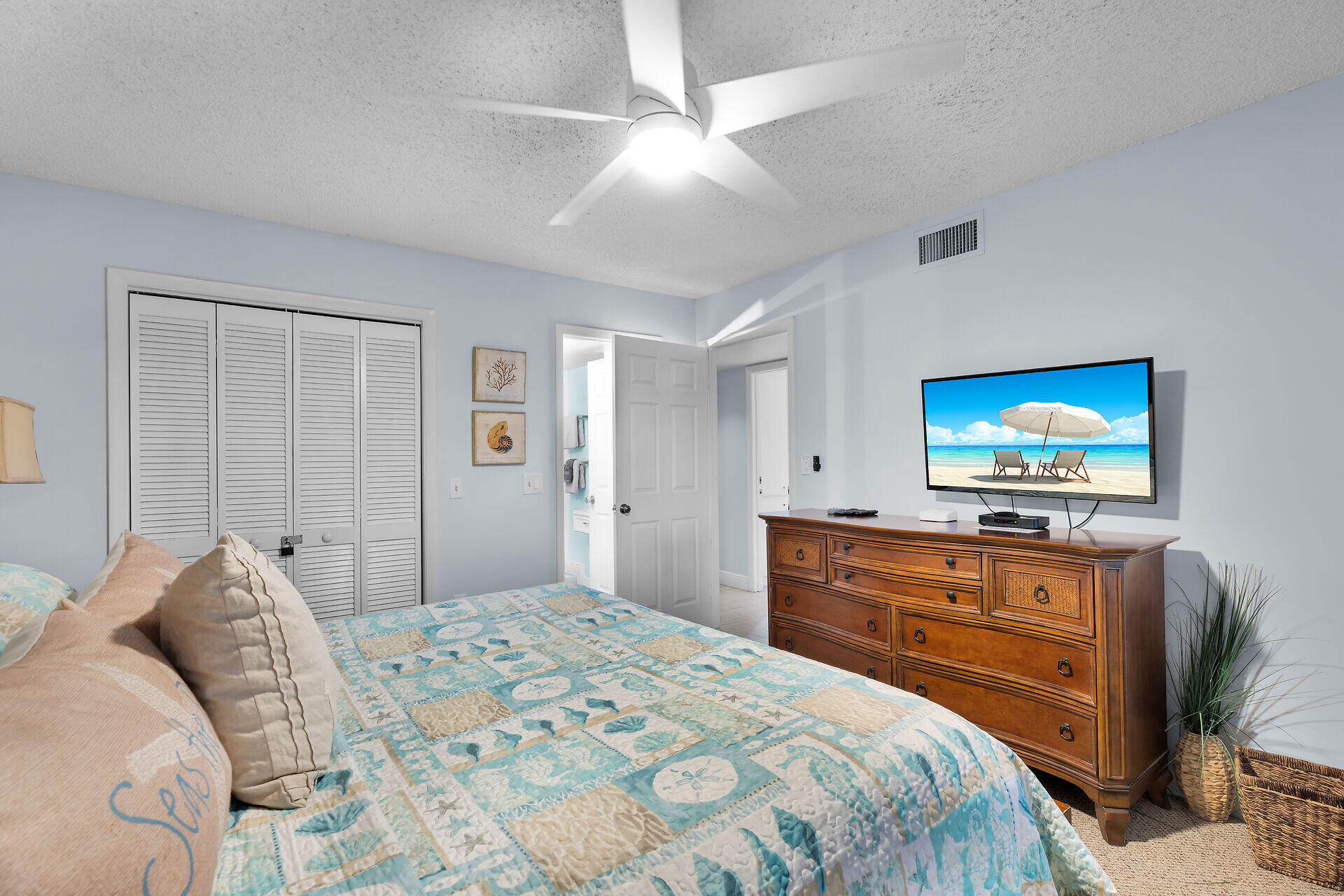 Furnished bedroom with a coastal-patterned quilt, ceiling fan, and wooden dresser with television.