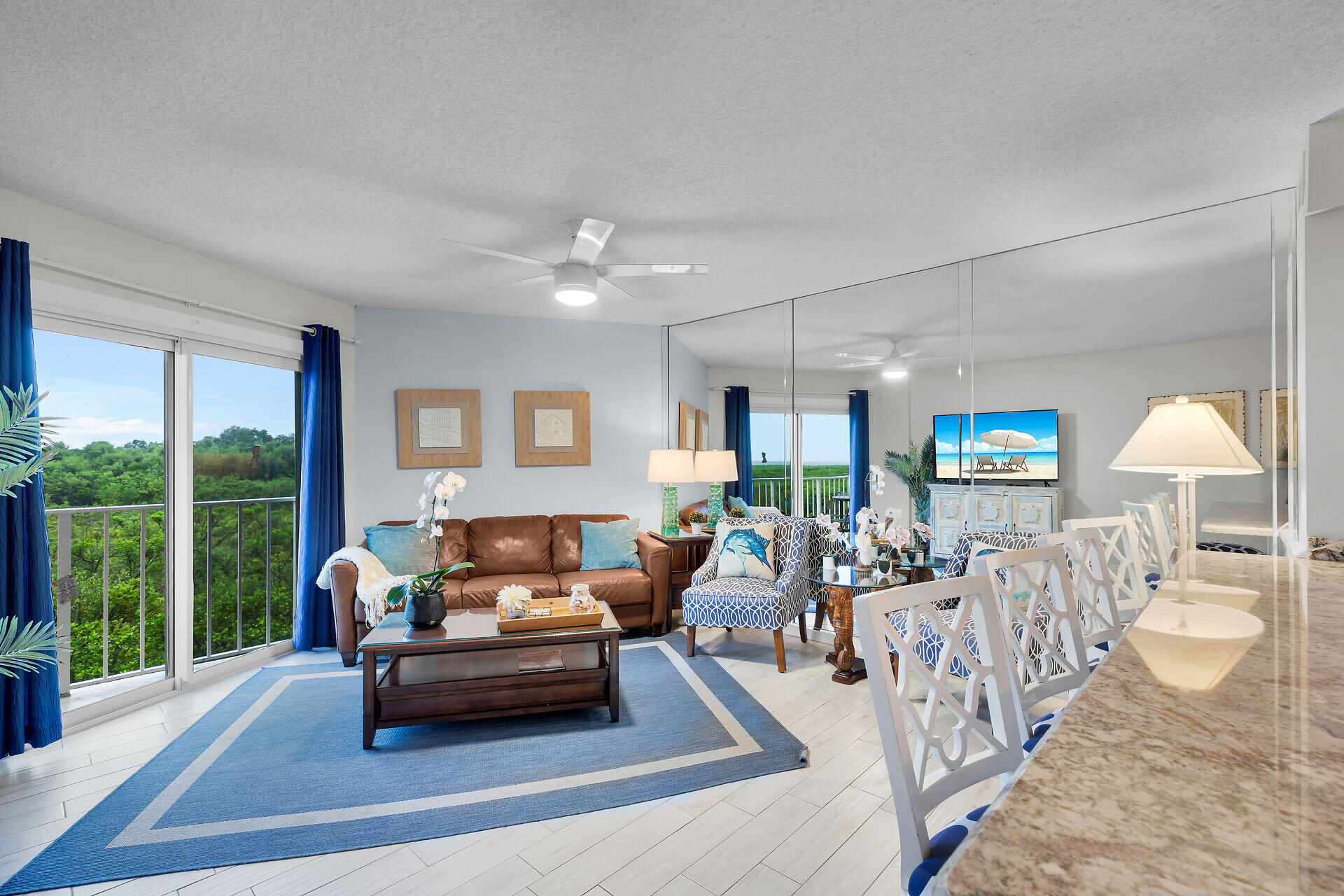 Furnished living and dining room with mirrored walls and a balcony overlooking greenery.