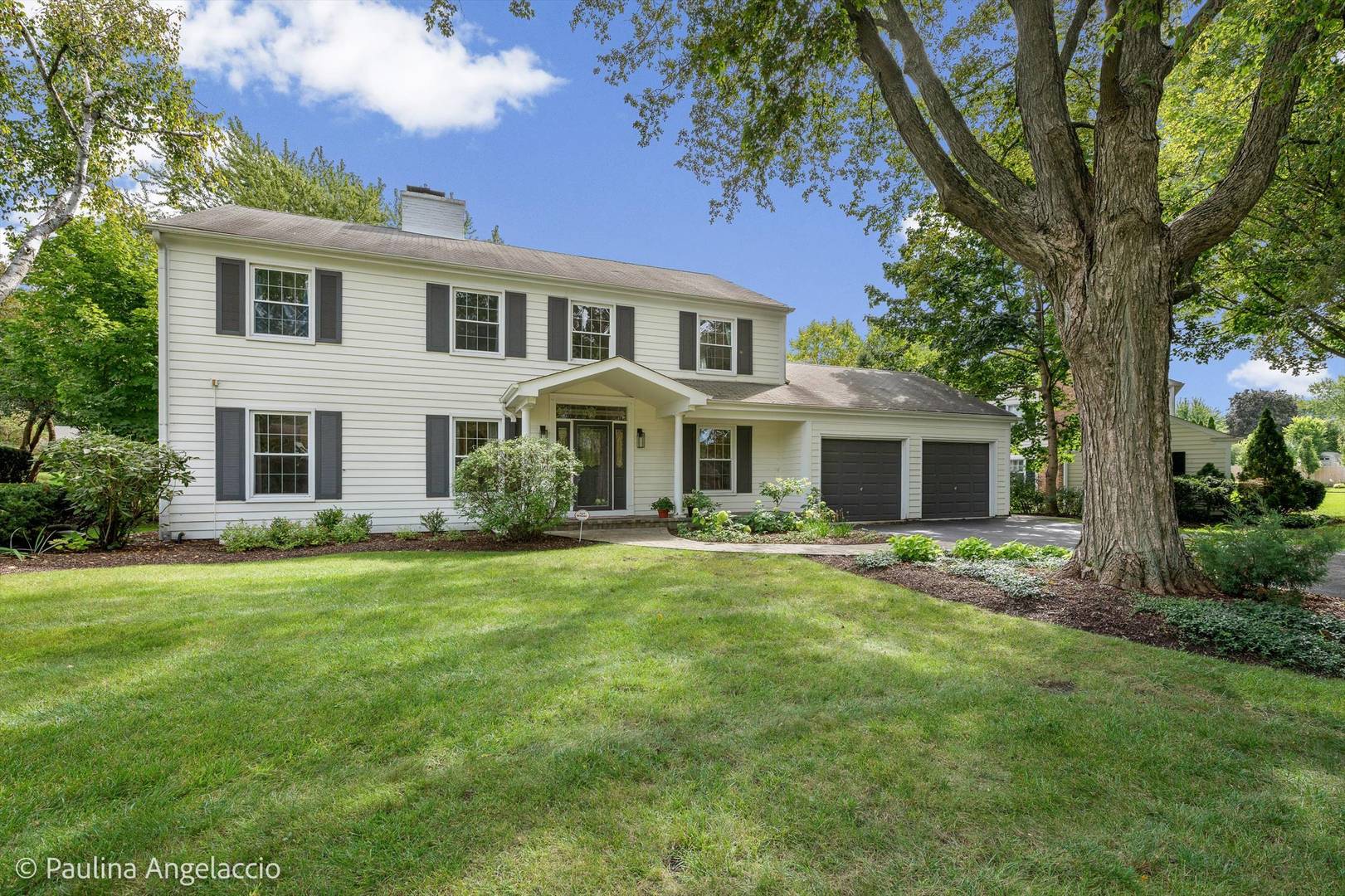 Two-story white house with dark shutters and a two-car garage beneath a large shade tree.