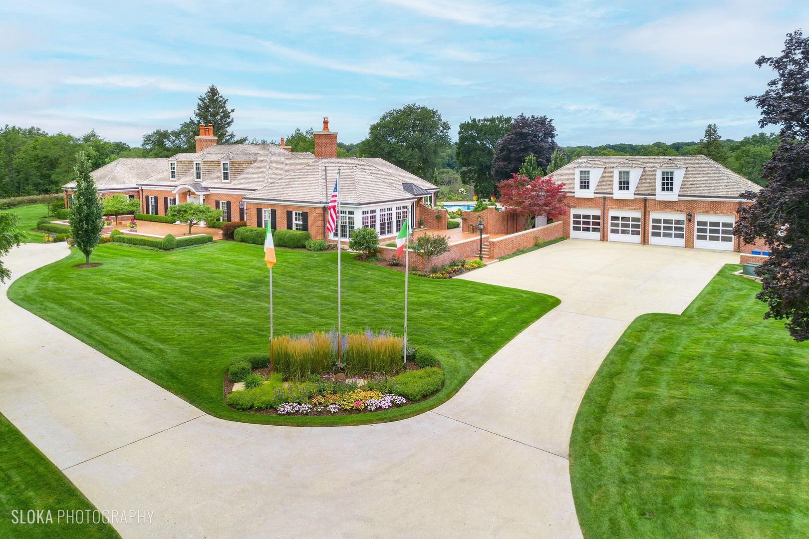 Large brick home and garage with a sweeping driveway, manicured lawns, and landscaped gardens.