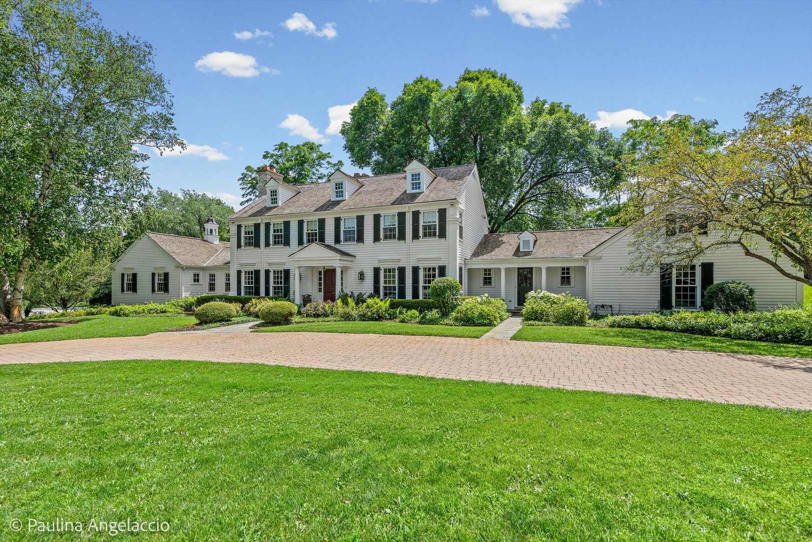 White two-story home with black shutters, landscaped gardens, and a broad curved brick driveway.